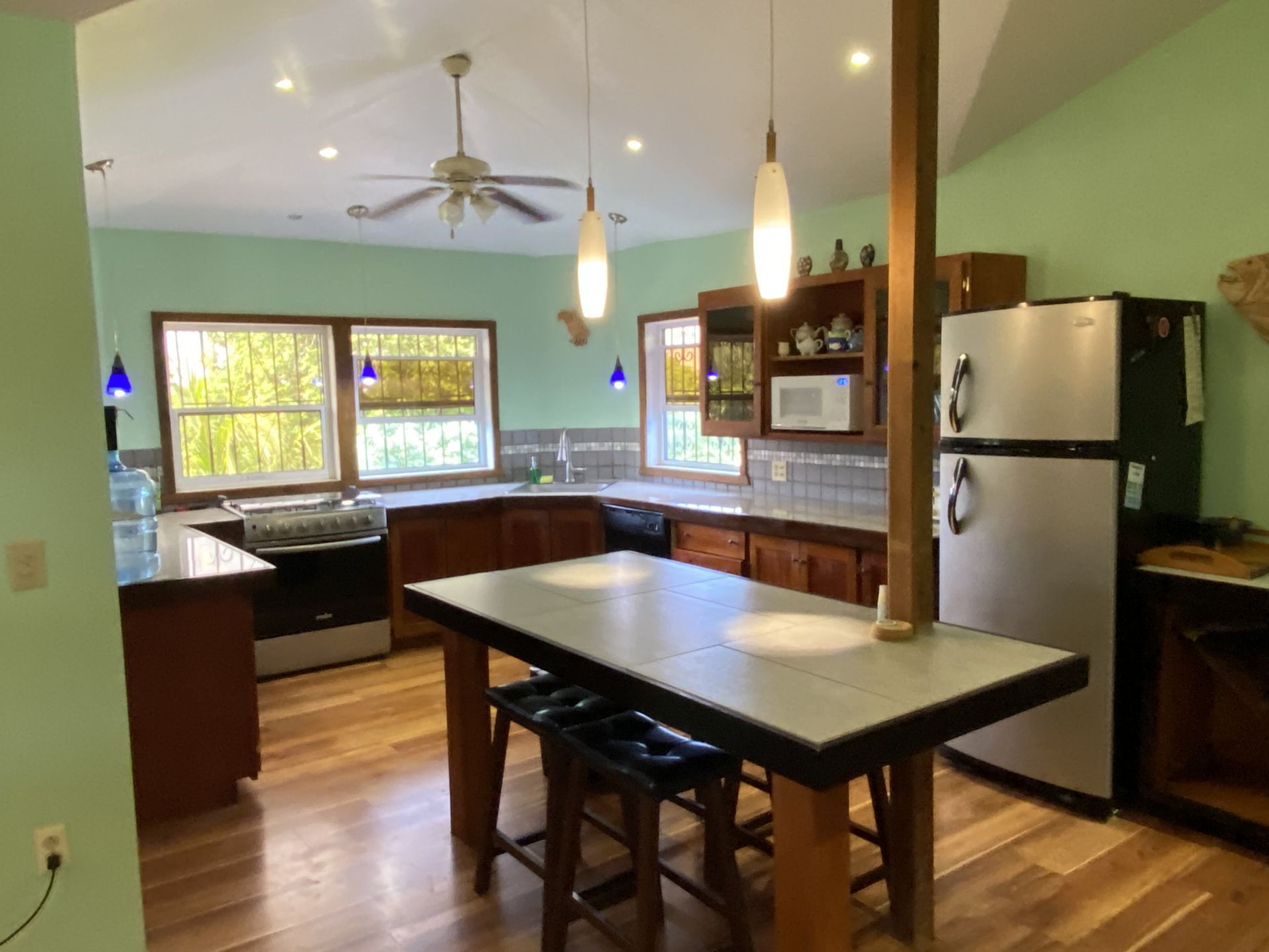 Kitchen with island, wooden cabinets, and light green walls; two pendant lights hang above island, windows in the background.