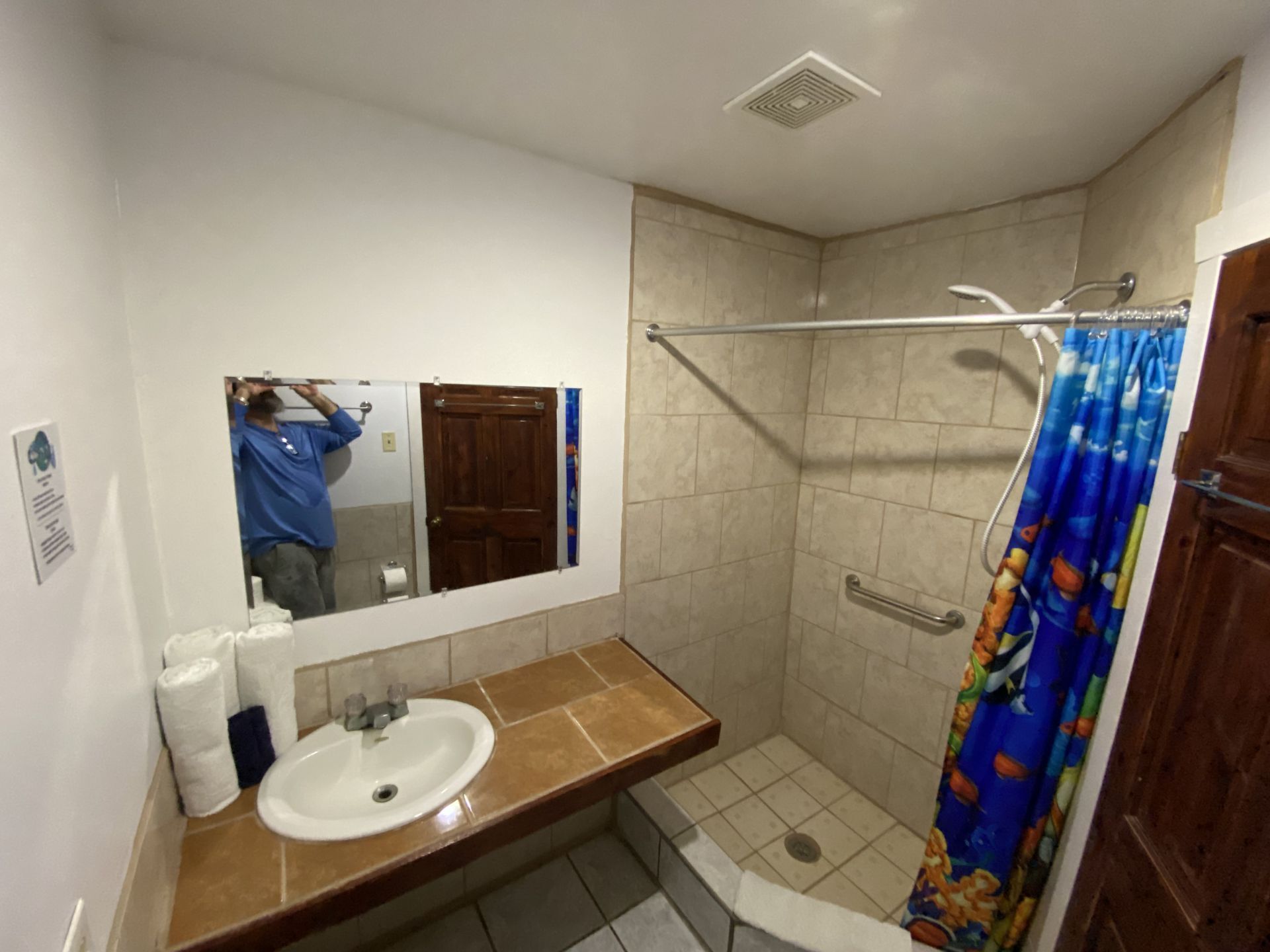 Bathroom with tan tiled shower, sink, mirror, and blue fish shower curtain.