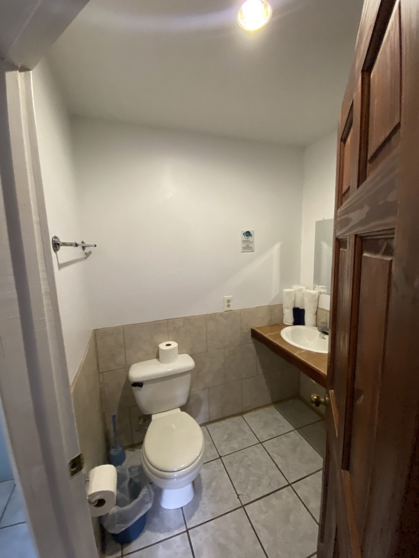 Small bathroom with toilet, sink, and a closed wooden door. White walls, beige tile, and a bright light.