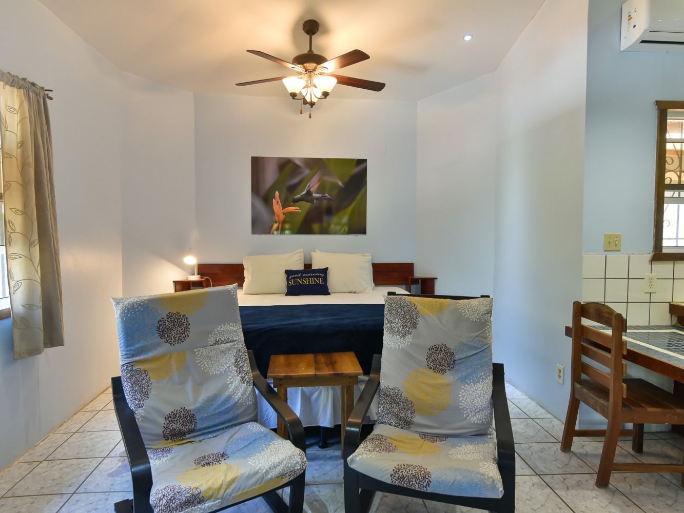 Bedroom with two chairs, bed, and artwork. White walls, tile floor.