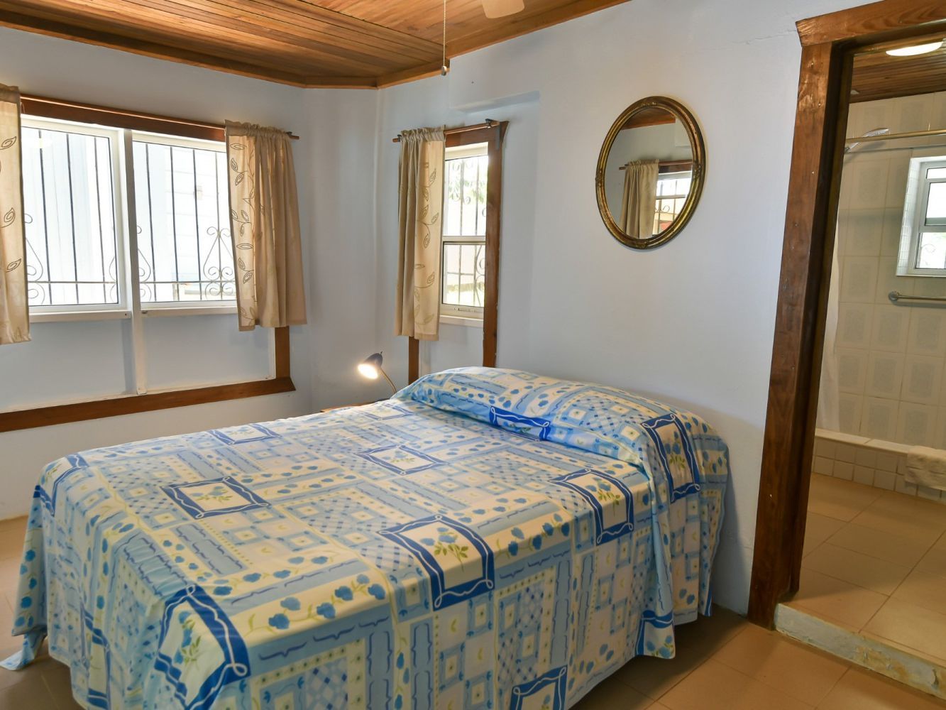 Bedroom with a double bed covered with a blue patterned blanket. A round mirror hangs on a white wall.