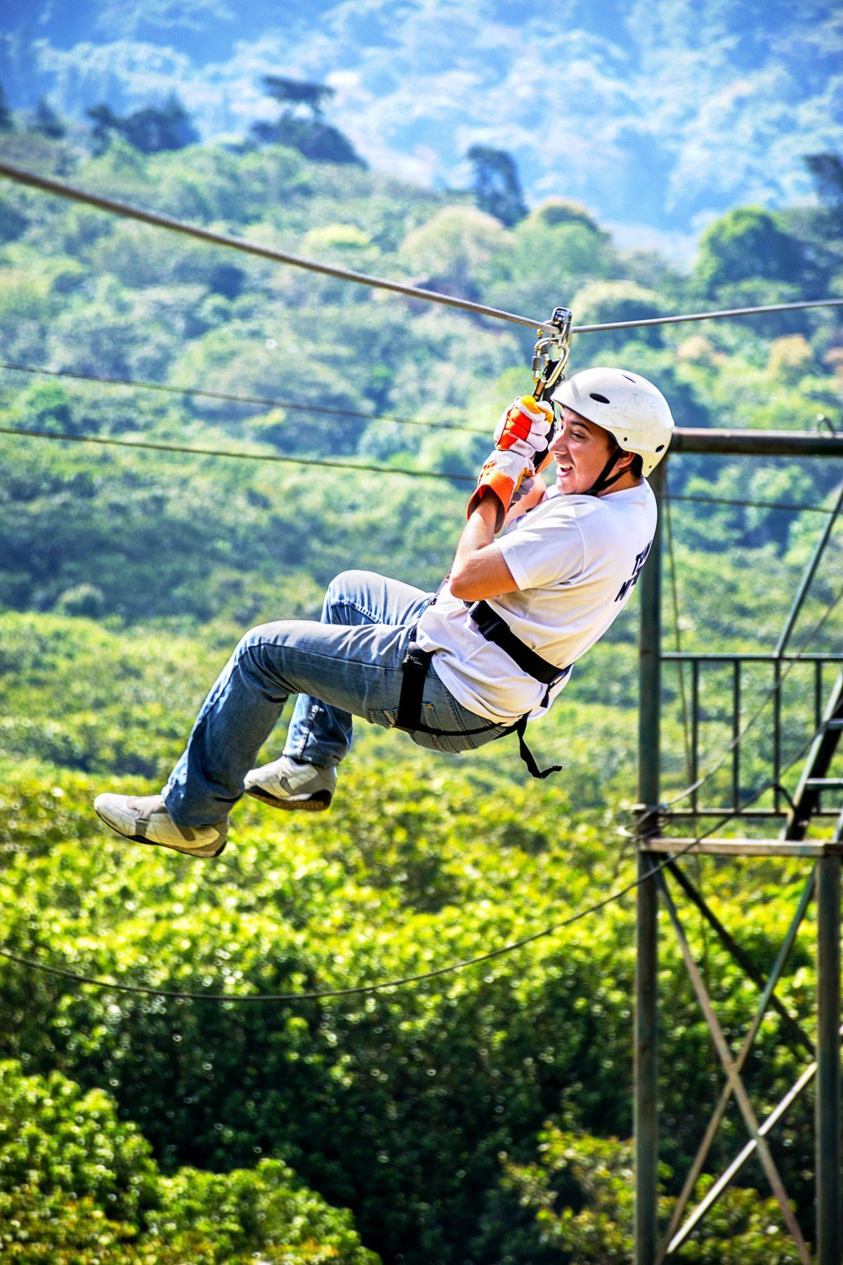 Person ziplining through a lush green forest, smiling, wearing a helmet and harness.