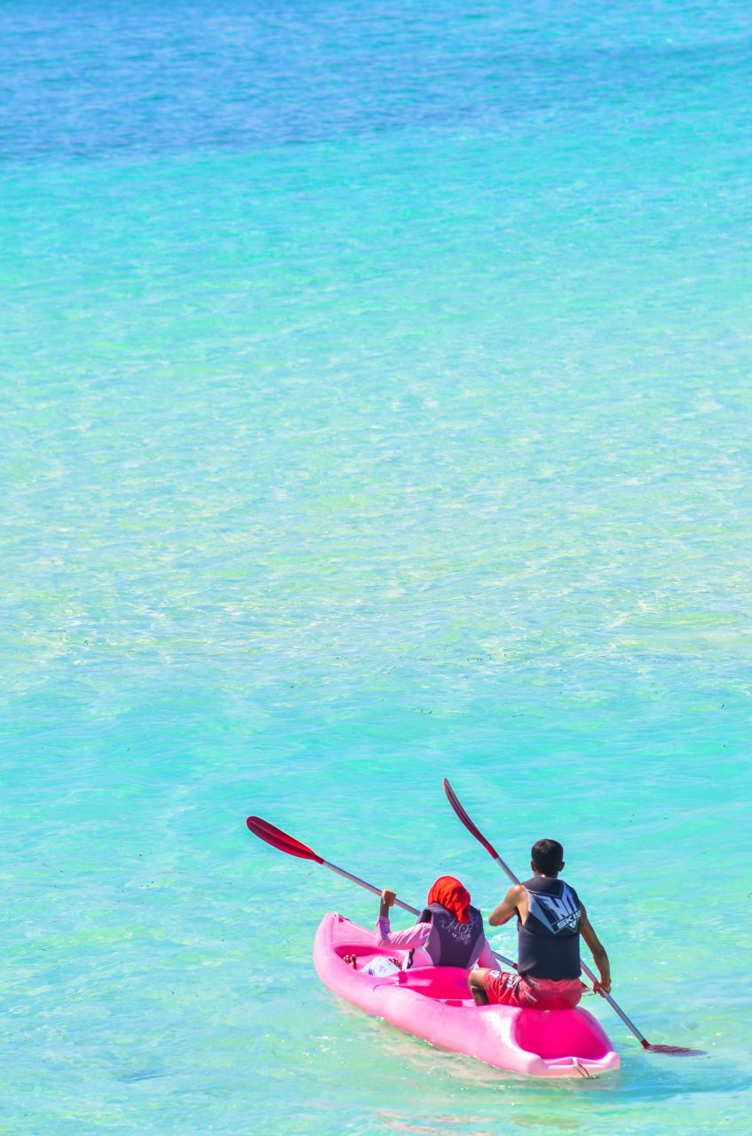 Two people kayaking in a pink kayak on clear turquoise water; sunny day.