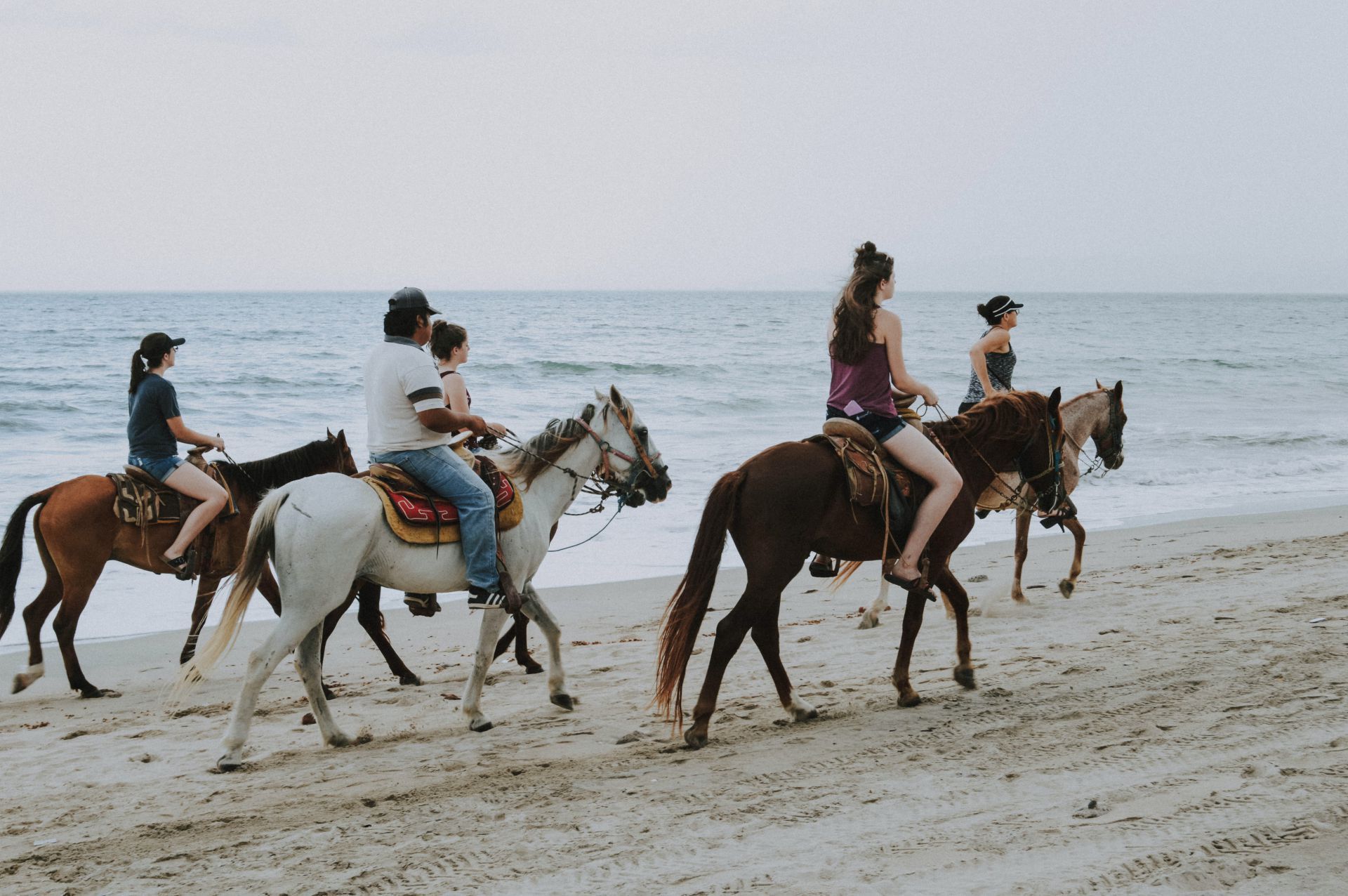 People riding horses on a sandy beach near the ocean, enjoying a leisurely ride on a cloudy day.
