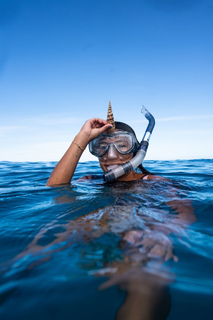 Woman snorkeling in ocean holds up a seashell, smiling, with blue water and sky.