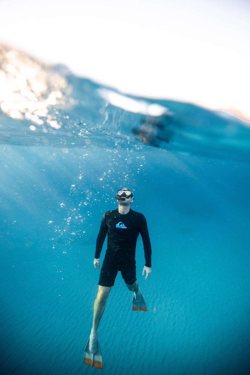 Man in wetsuit swims underwater with fins, bubbles, and the water's surface visible.