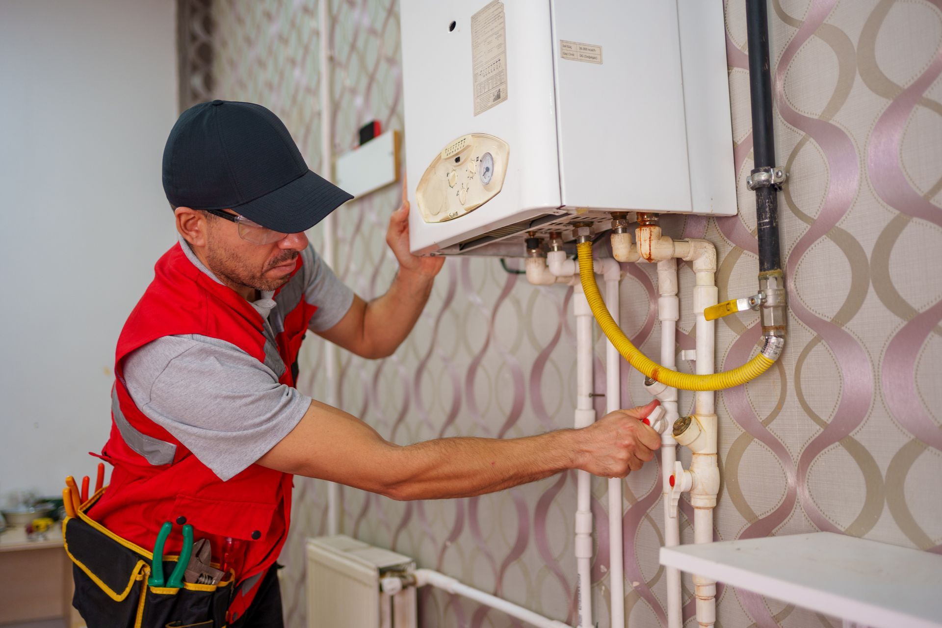 A technician wearing a red vest and hat carefully works on a residential heater installation. A technician wearing a red vest and hat carefully works on a residential heater installation.
