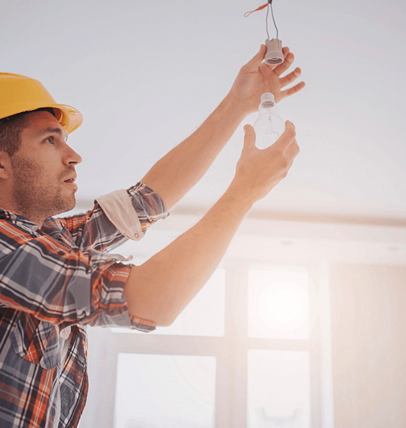 A Man in a Hard Hat is Hanging a Light Bulb From the Ceiling — Clifford Electrical in Lennox Head, NSW