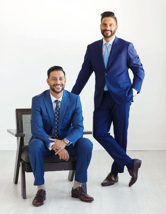 Two men in suits are standing next to each other . one is sitting in a chair and the other is standing.