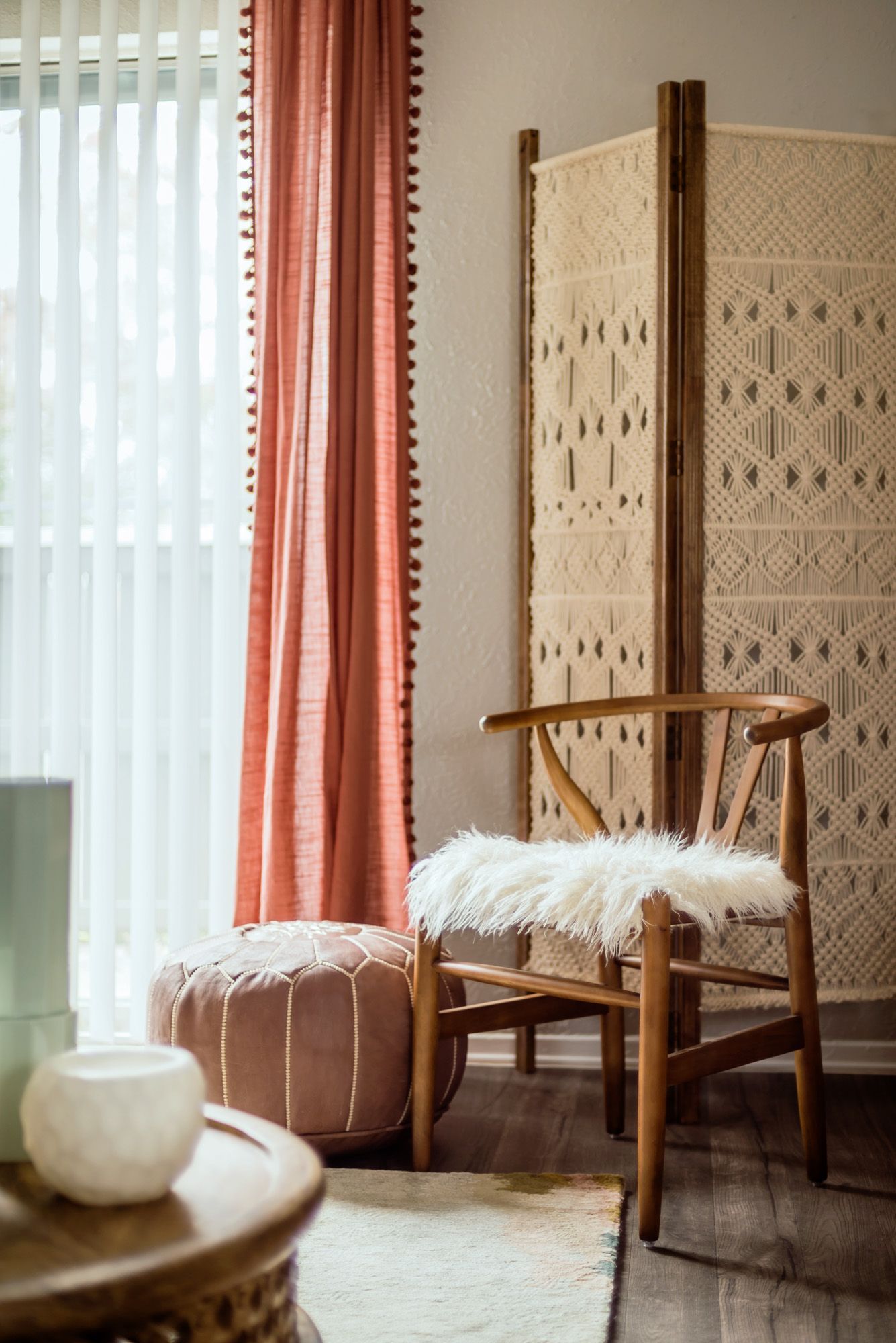 Boho-style room: pink curtains, wooden chair with fur, macrame screen, pink ottoman, and small table.