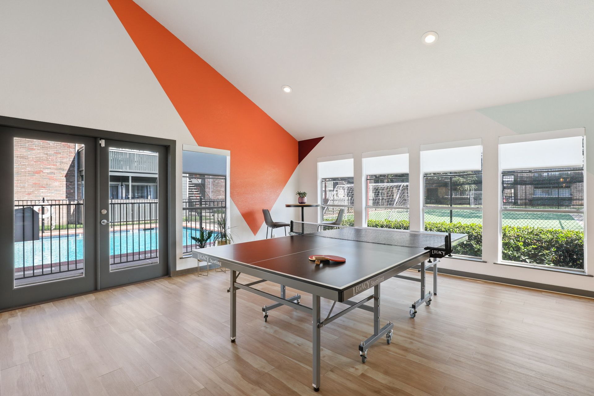 Ping pong table in a room with large windows overlooking a pool and an orange wall design.