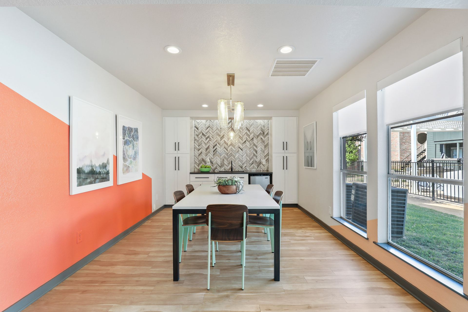 Long dining table in a room with orange and white walls, windows, and light wood flooring.
