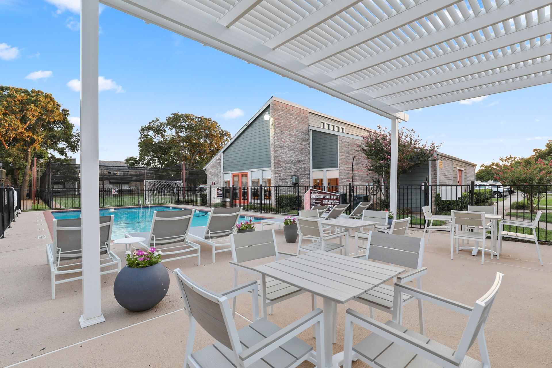 Patio area with tables, chairs, and a swimming pool, beneath a pergola. Sunny day.