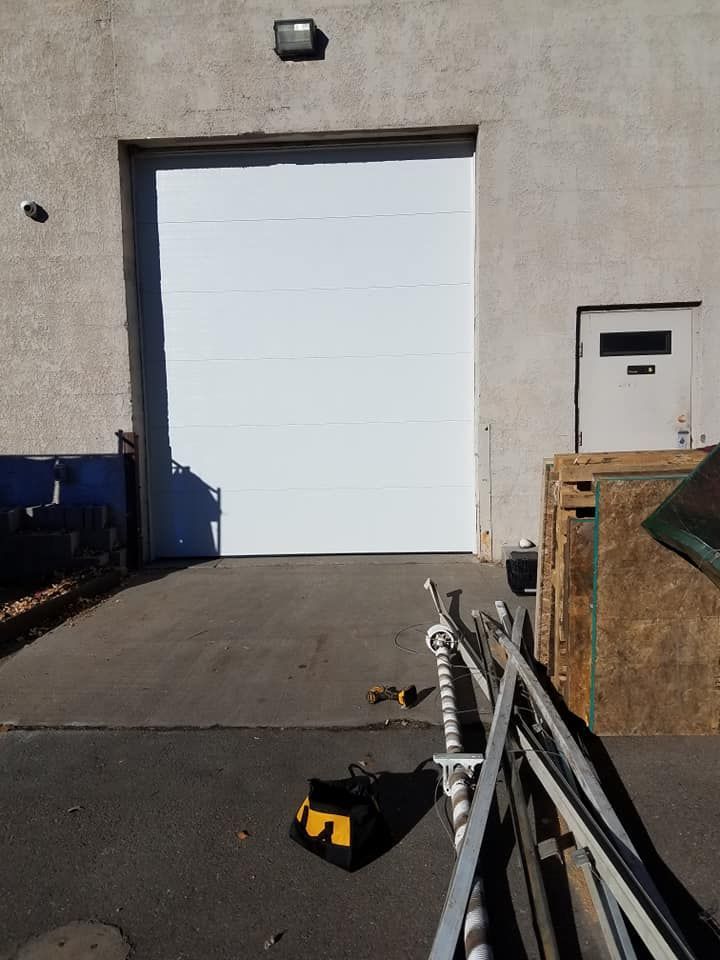 A white industrial bay door on a concrete building, with construction materials and tools on the ground nearby.