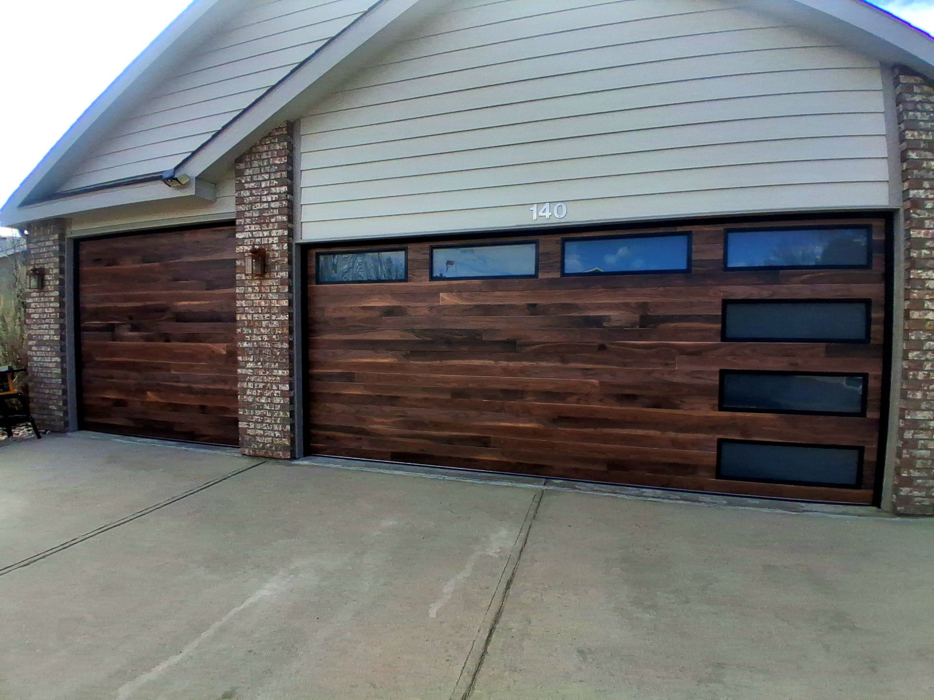 Two dark wood garage doors with rectangular glass windows on a house with beige siding and brick pillars.