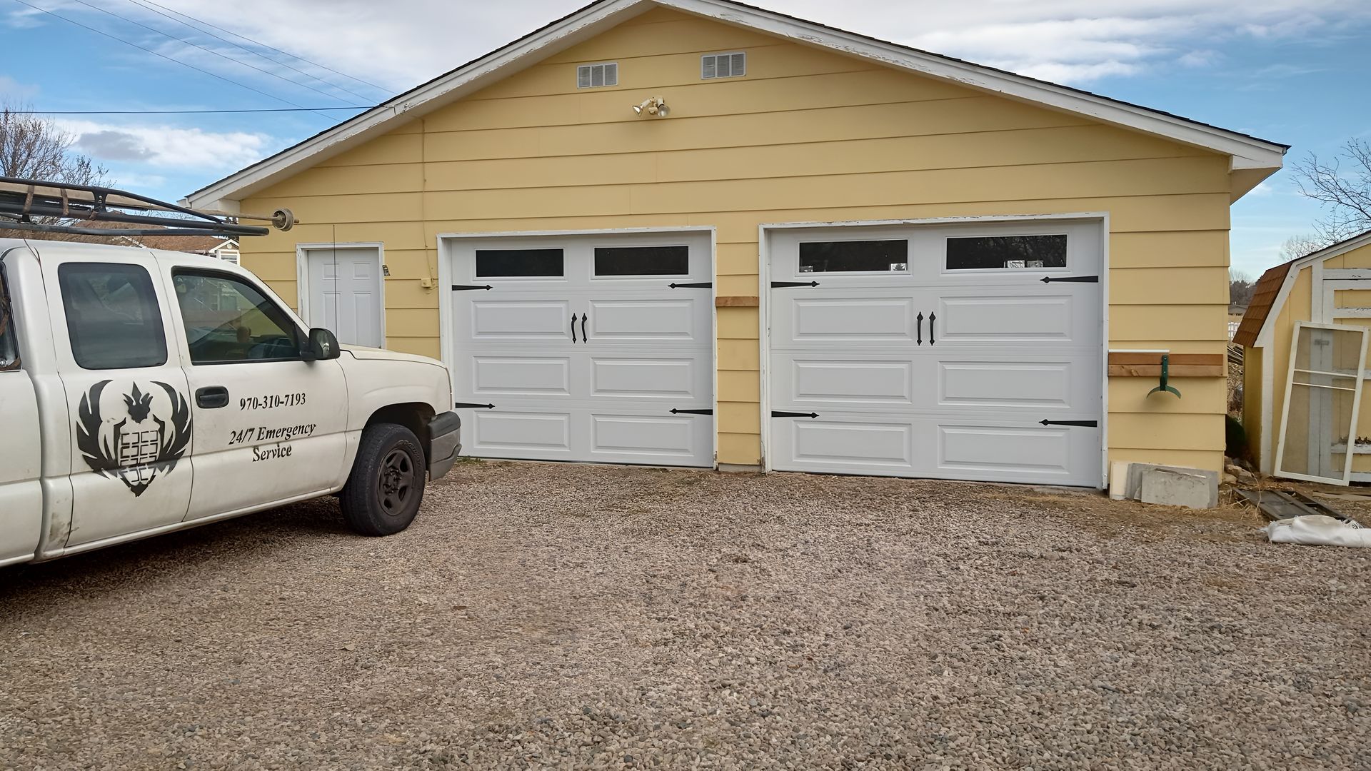 A white pickup truck parked in front of a yellow garage with two white bay doors under a partially cloudy sky.