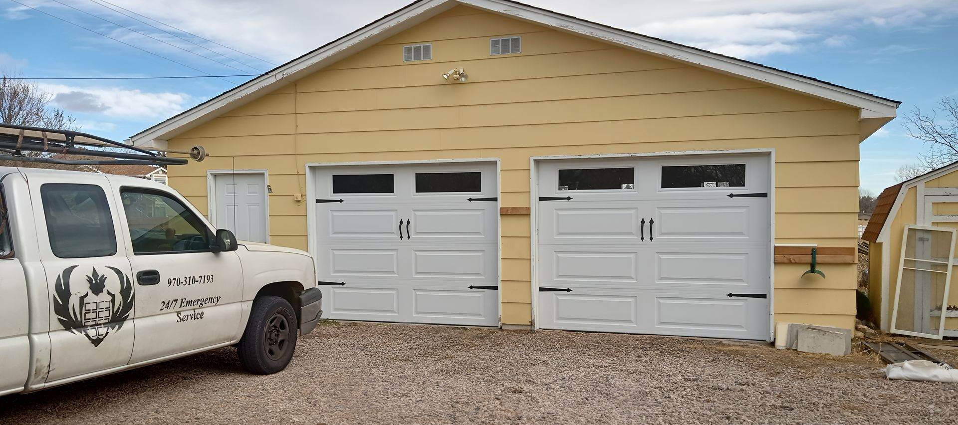 A yellow garage with two white doors sits next to a white service truck on a gravel lot under a cloudy sky.