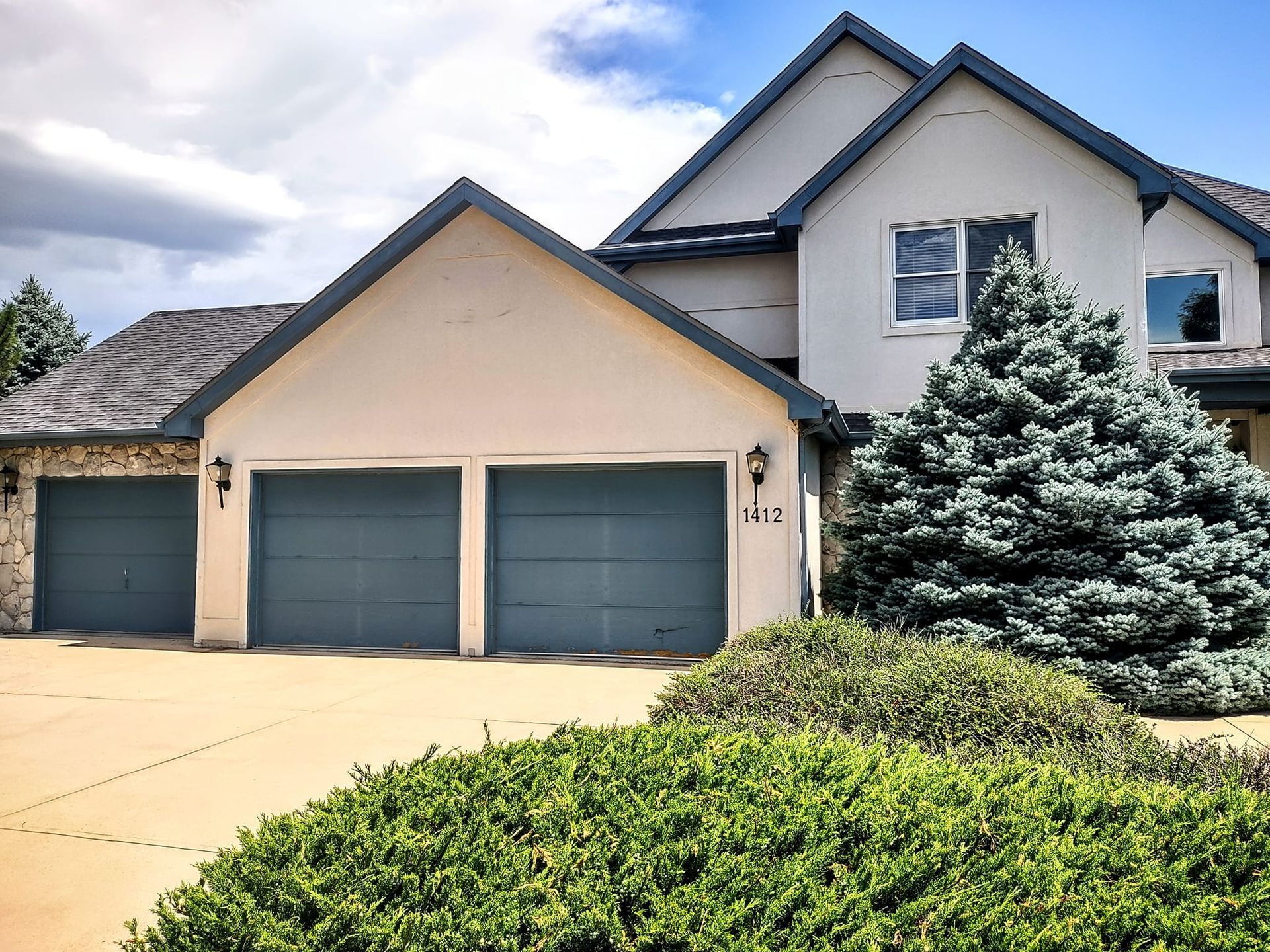 A multi-level home with three blue garage doors, beige stucco walls, and a large evergreen bush in the front yard.