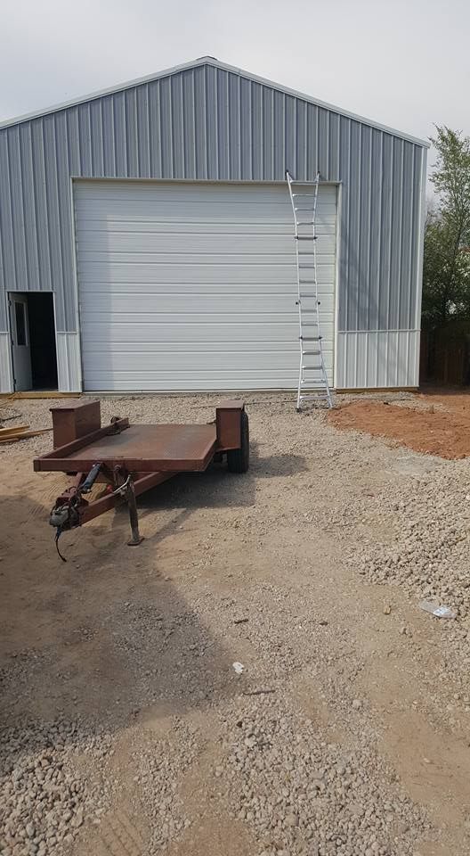 A tall metal-sided building with a large white roll-up door and a ladder leaning against it, parked before a trailer.