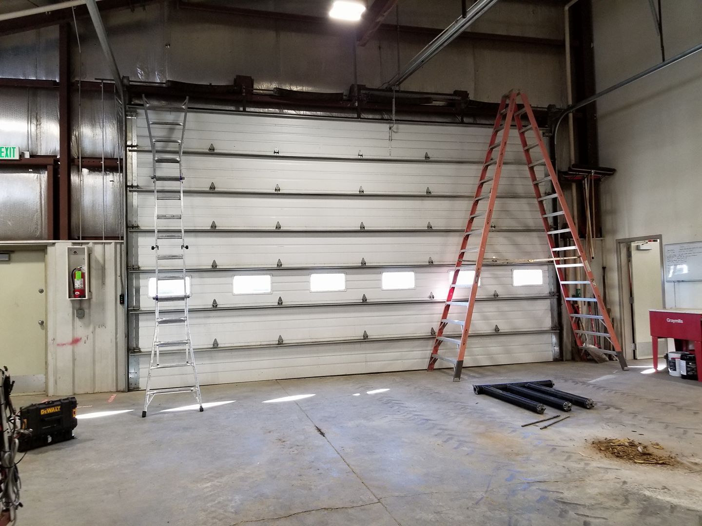 Large industrial space with a closed white sectional garage door, two ladders, and tools on the concrete floor.