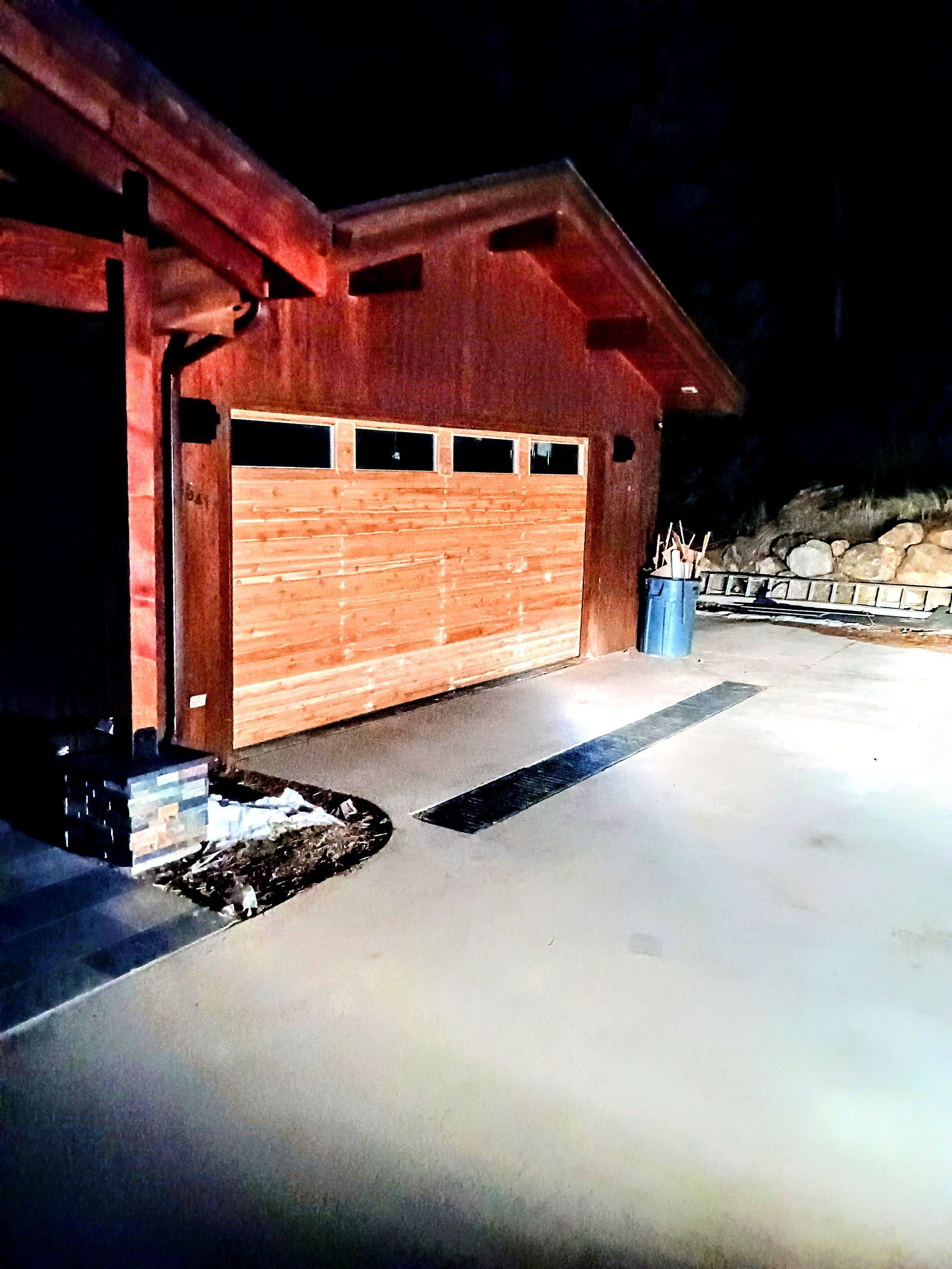 A wooden garage with horizontal panels and rectangular windows, lit at night with a paved driveway in the foreground.