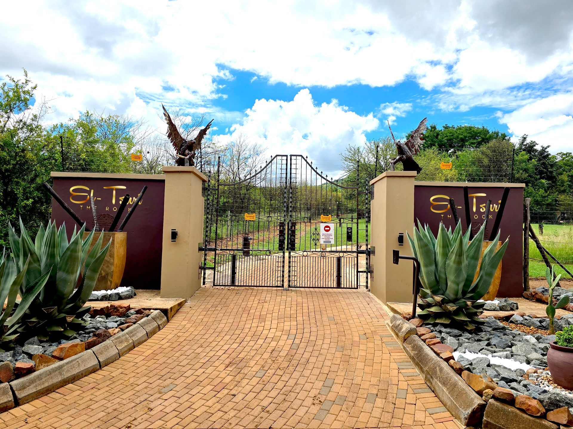 Brick pathway leading to gated entrance with eagle statues and landscaping under a blue sky.