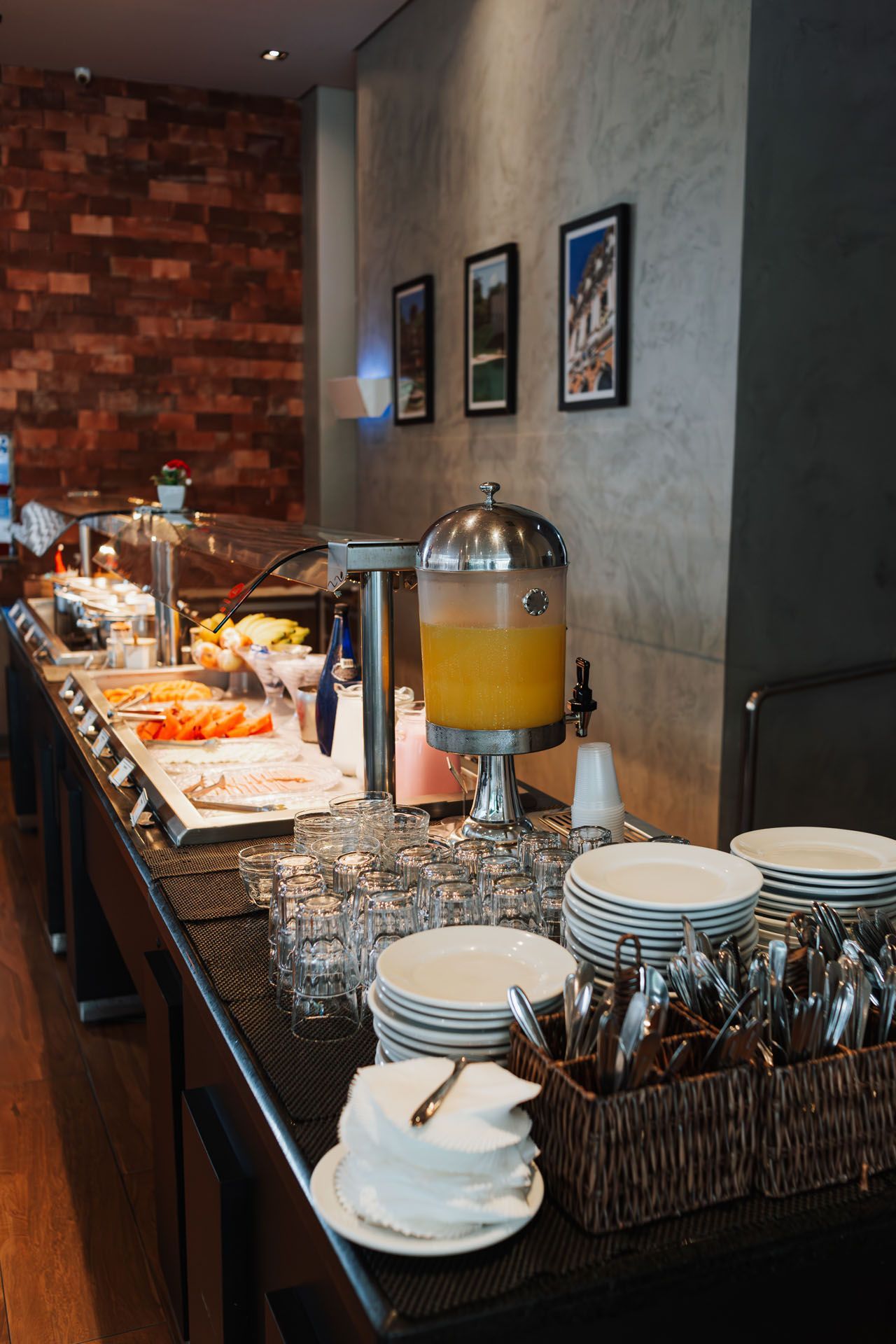Mesa de buffet com frutas, dispensador de suco, pratos e talheres em um restaurante.