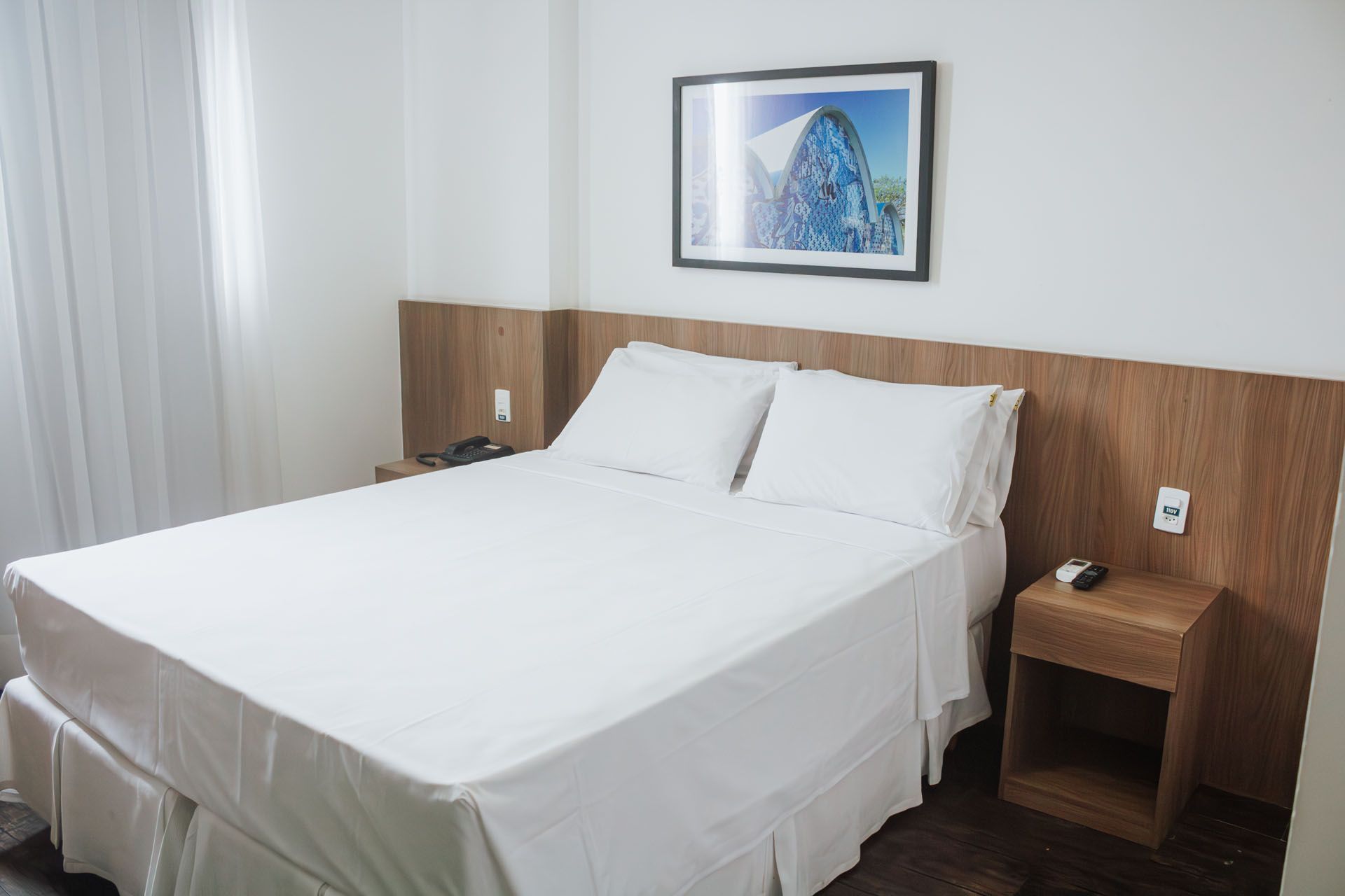Quarto de hotel com cama branca, cabeceira e mesa de cabeceira de madeira, e obras de arte na parede.