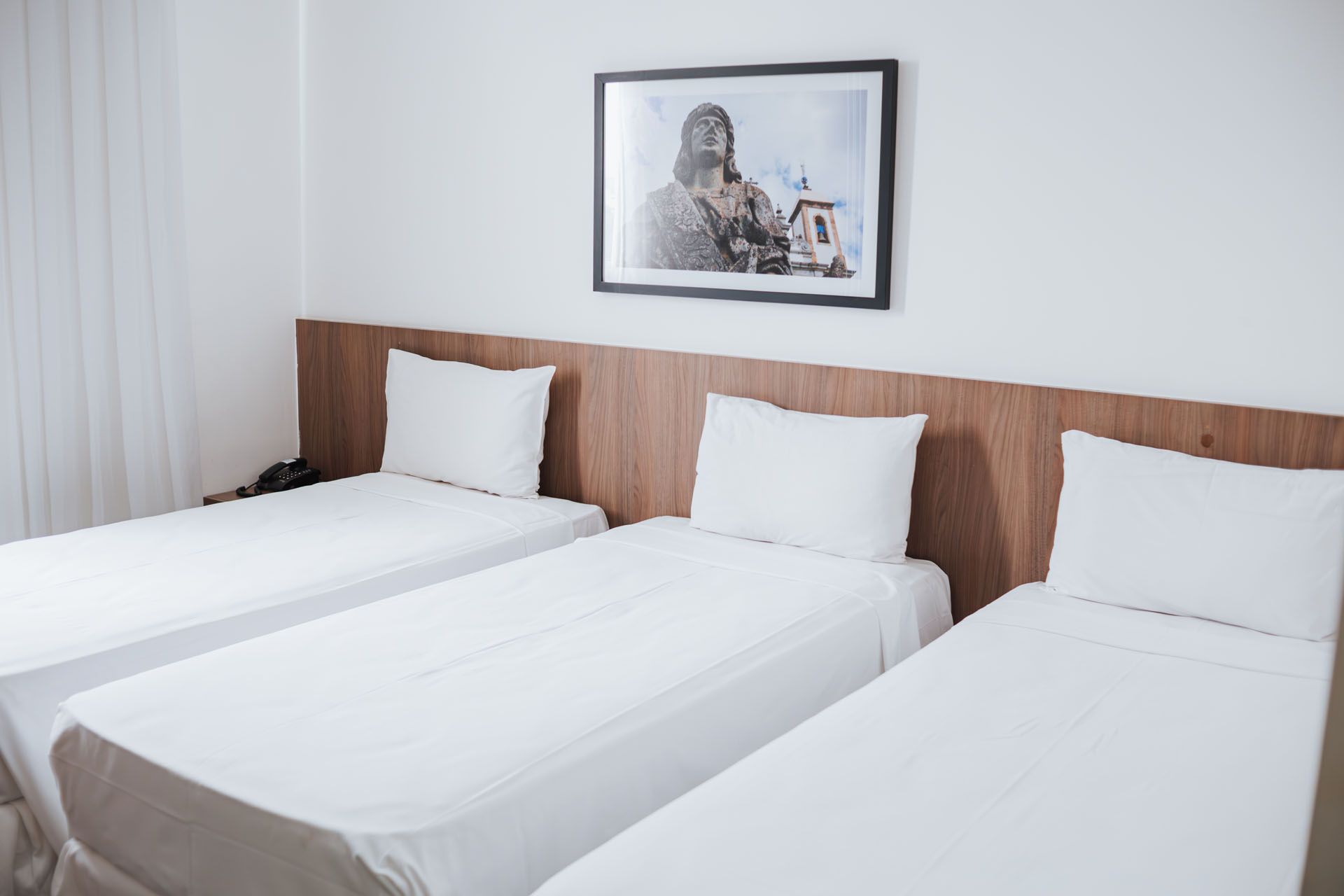 Três camas de solteiro em um quarto de hotel com lençóis brancos, cabeceira de madeira e um quadro na parede.