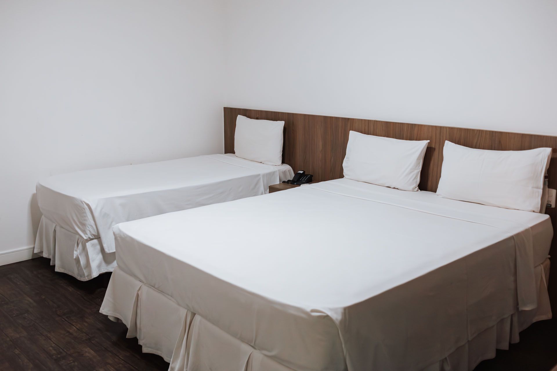 Quarto de hotel com uma cama de casal e uma cama de solteiro; roupa de cama branca, cabeceira de madeira, piso escuro.