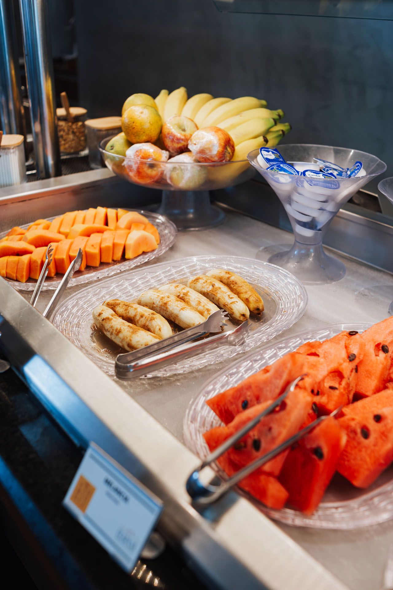 Buffet de frutas: mamão papaia, banana frita, melancia e uma tigela de vidro com frutas variadas.