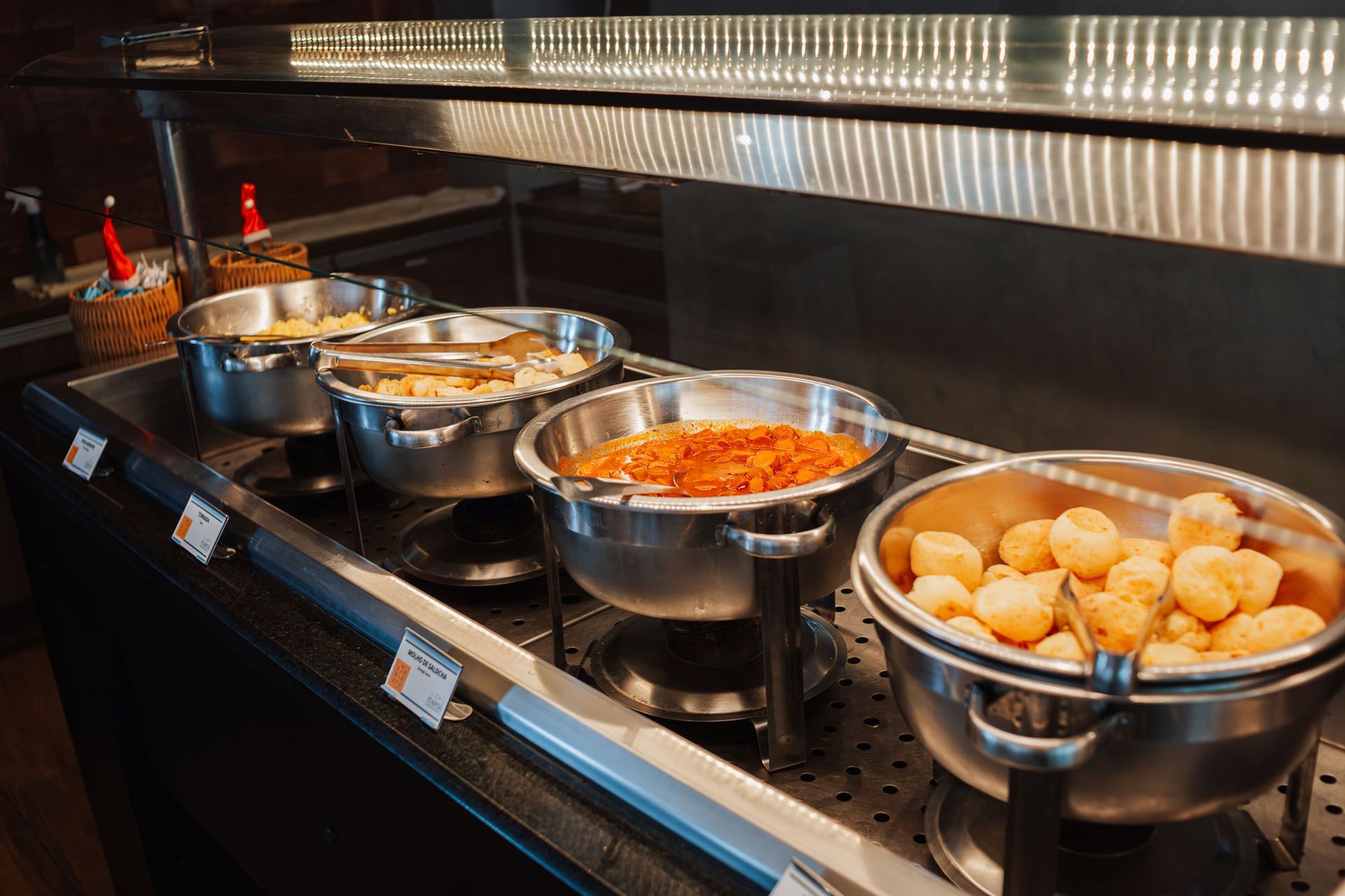 Buffet de comida com vários recipientes de aço inoxidável contendo diferentes pratos, iluminados por cima.