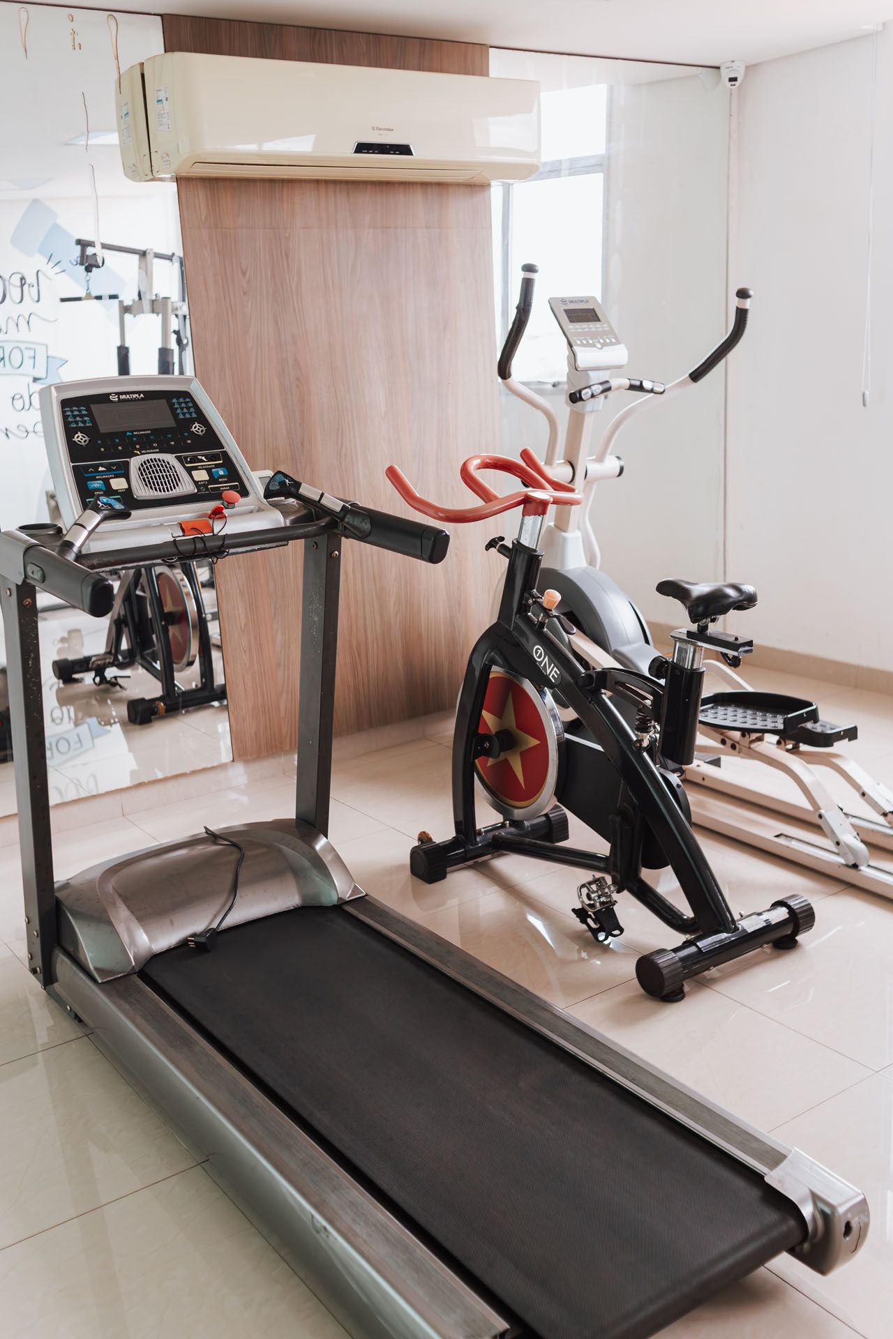 Esteira e bicicleta ergométrica em uma academia, paredes bege e piso de azulejo branco.