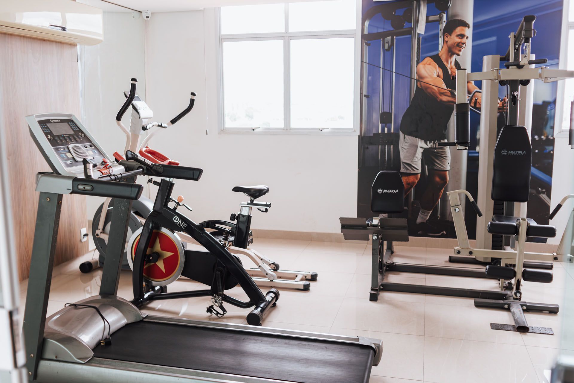 Interior de academia com esteira, bicicletas ergométricas, aparelhos de musculação e um pôster motivacional.