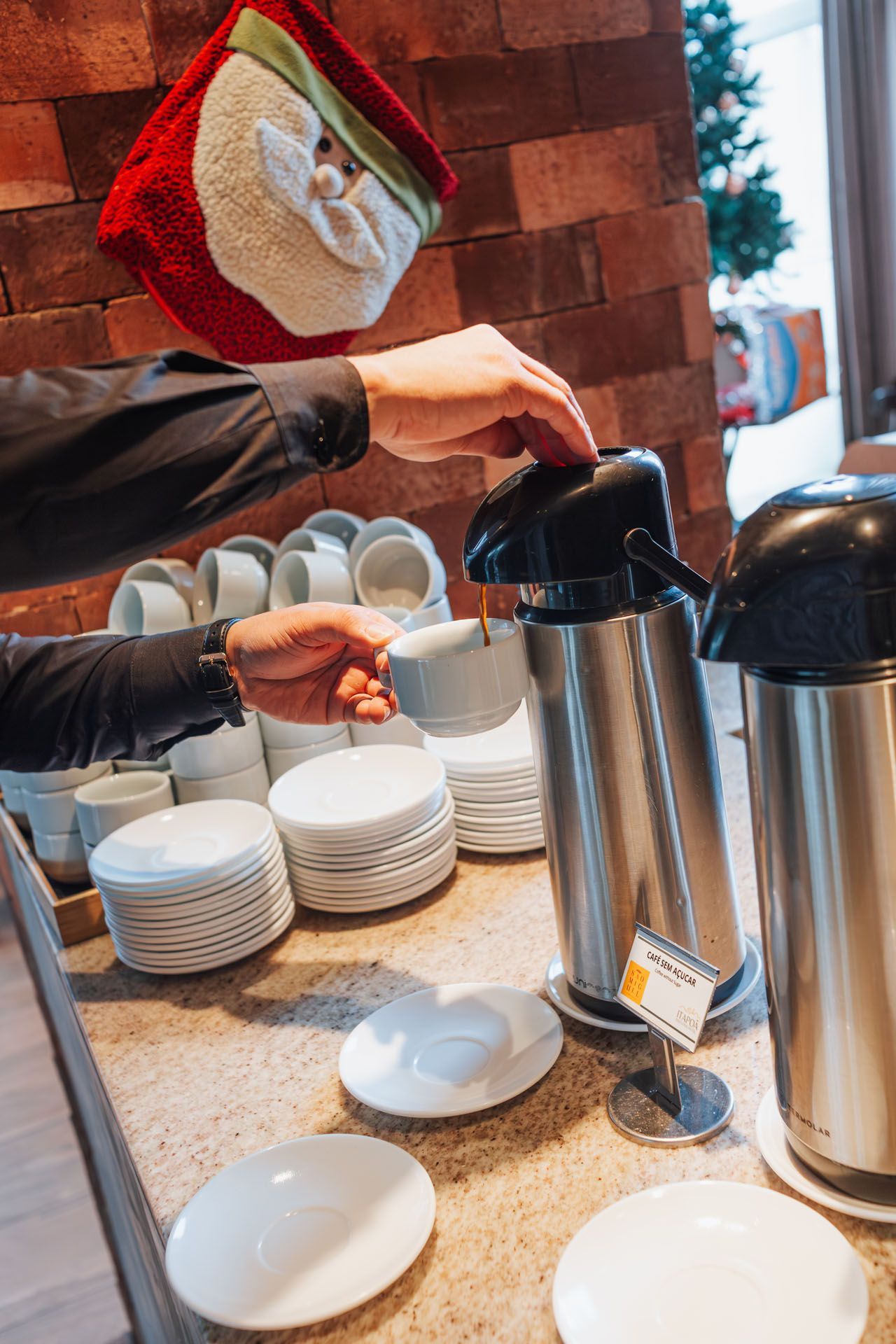 Uma pessoa serve café de uma garrafa térmica prateada para uma caneca branca em um aparador.