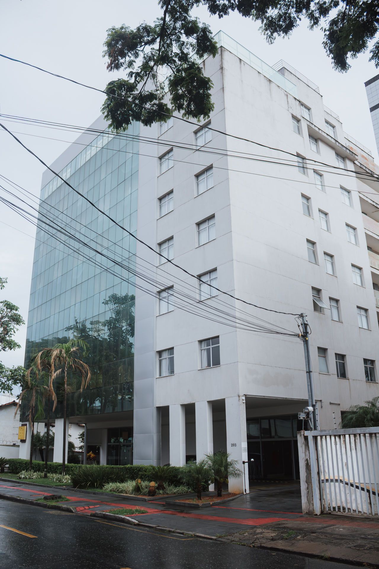 Prédio de escritórios branco com fachada de vidro, ao lado de uma rua com fiação elétrica, sob céu nublado.