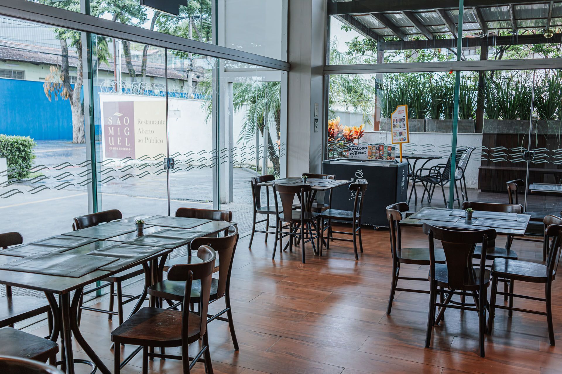 Interior vazio de um restaurante com mesas, cadeiras e grandes janelas com vista para o exterior.
