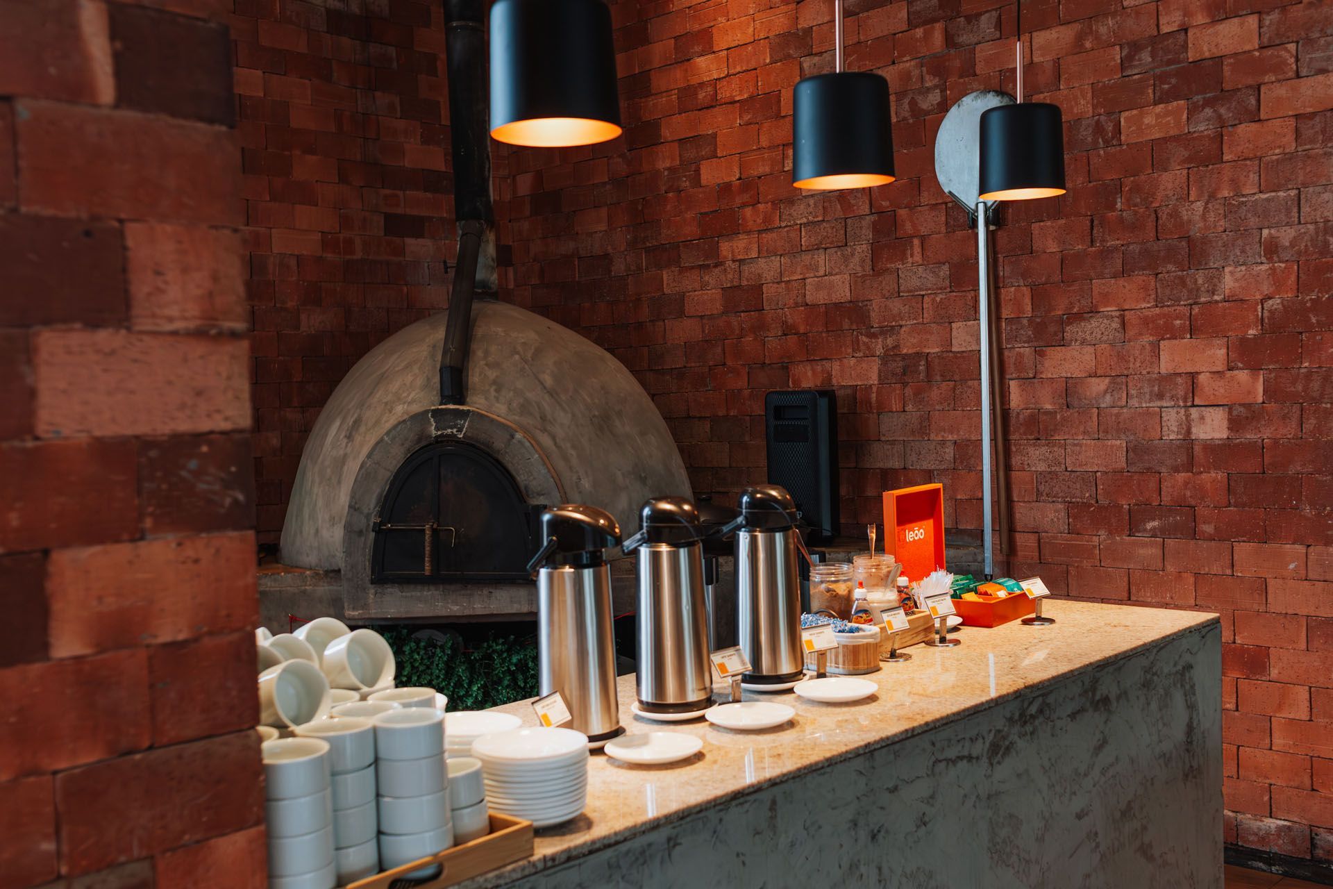 Interior com paredes de tijolos, forno de pizza, espaço para café e luminárias pendentes pretas.