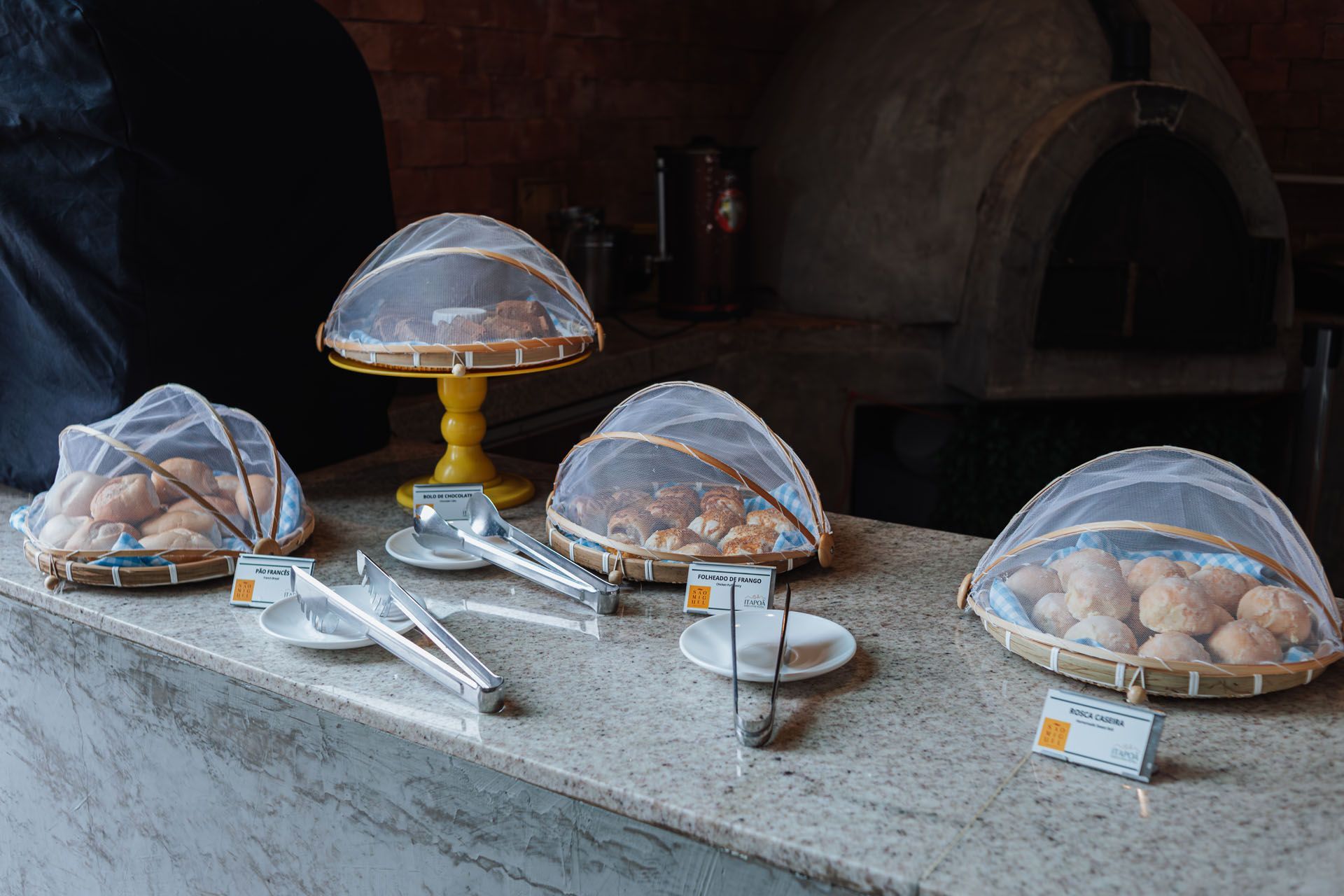 Expositor de pães com cestas cobertas sobre uma bancada, perto de um forno de tijolos. Pinças de prata e pratinhos também estão presentes.