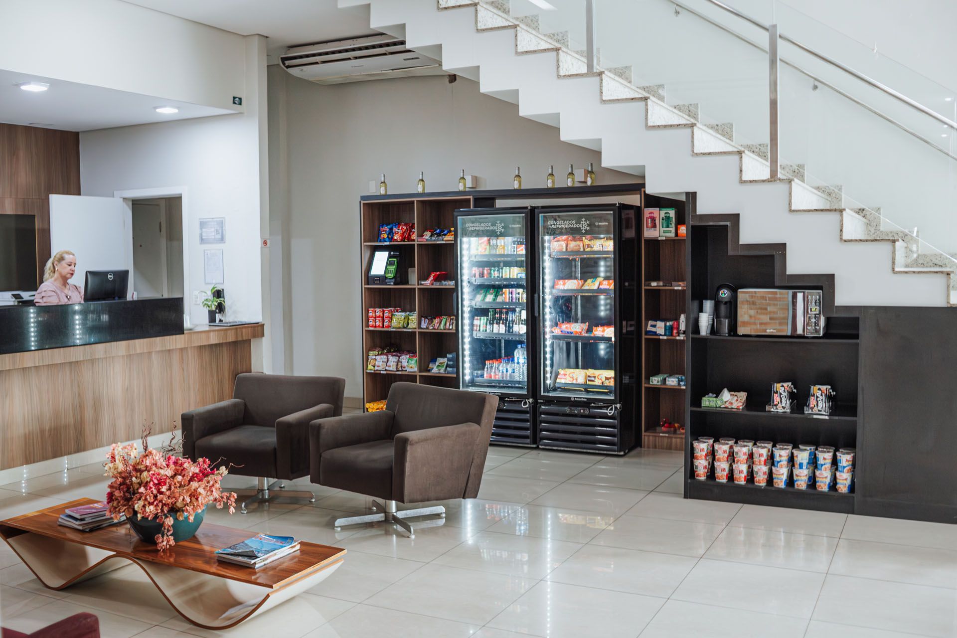 Hall de entrada do hotel com balcão de recepção, área de estar e expositor de lanches/bebidas. Escadas levam ao andar superior.