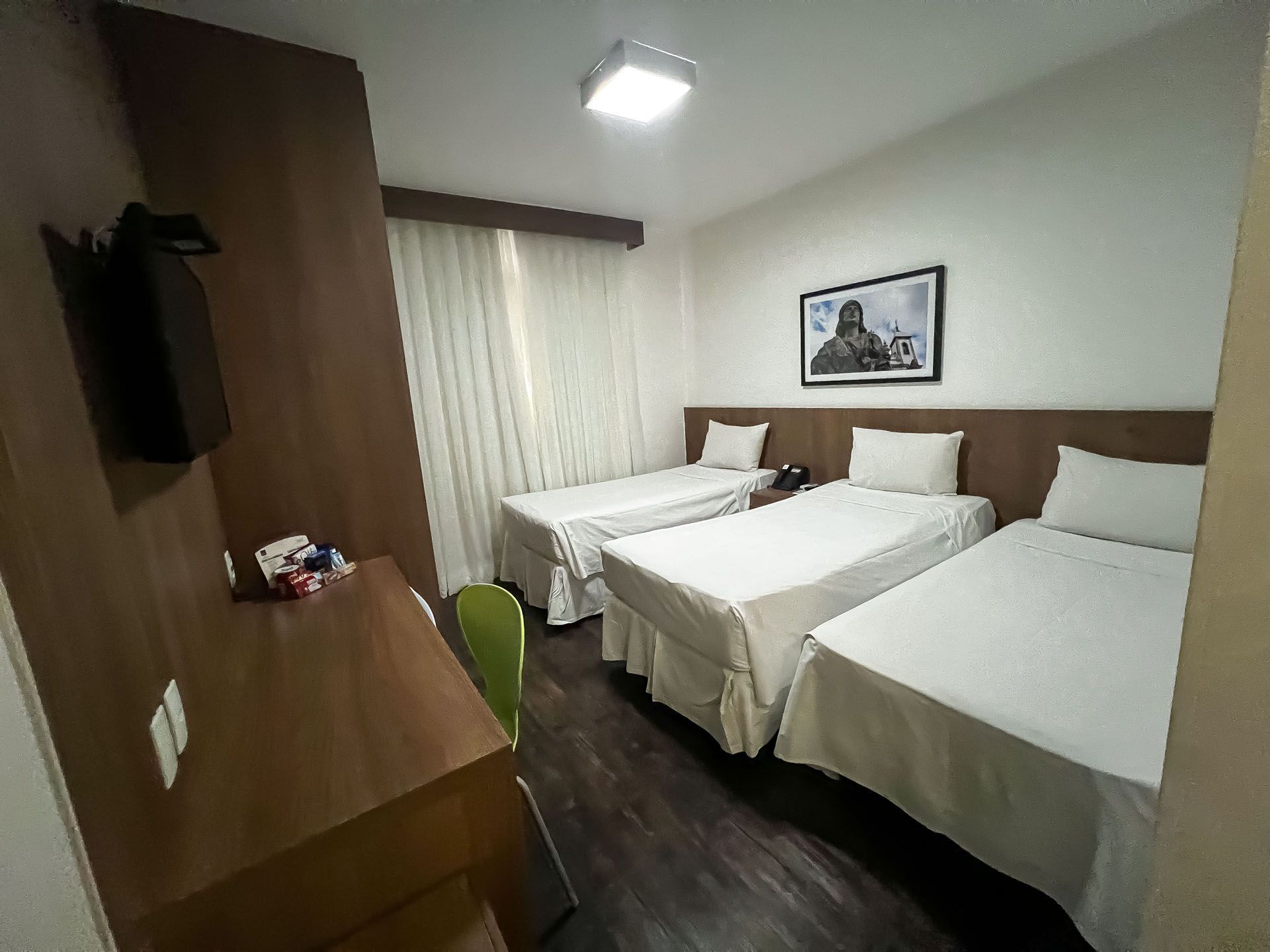 Quarto de hotel com três camas, uma escrivaninha e uma TV fixada na parede.