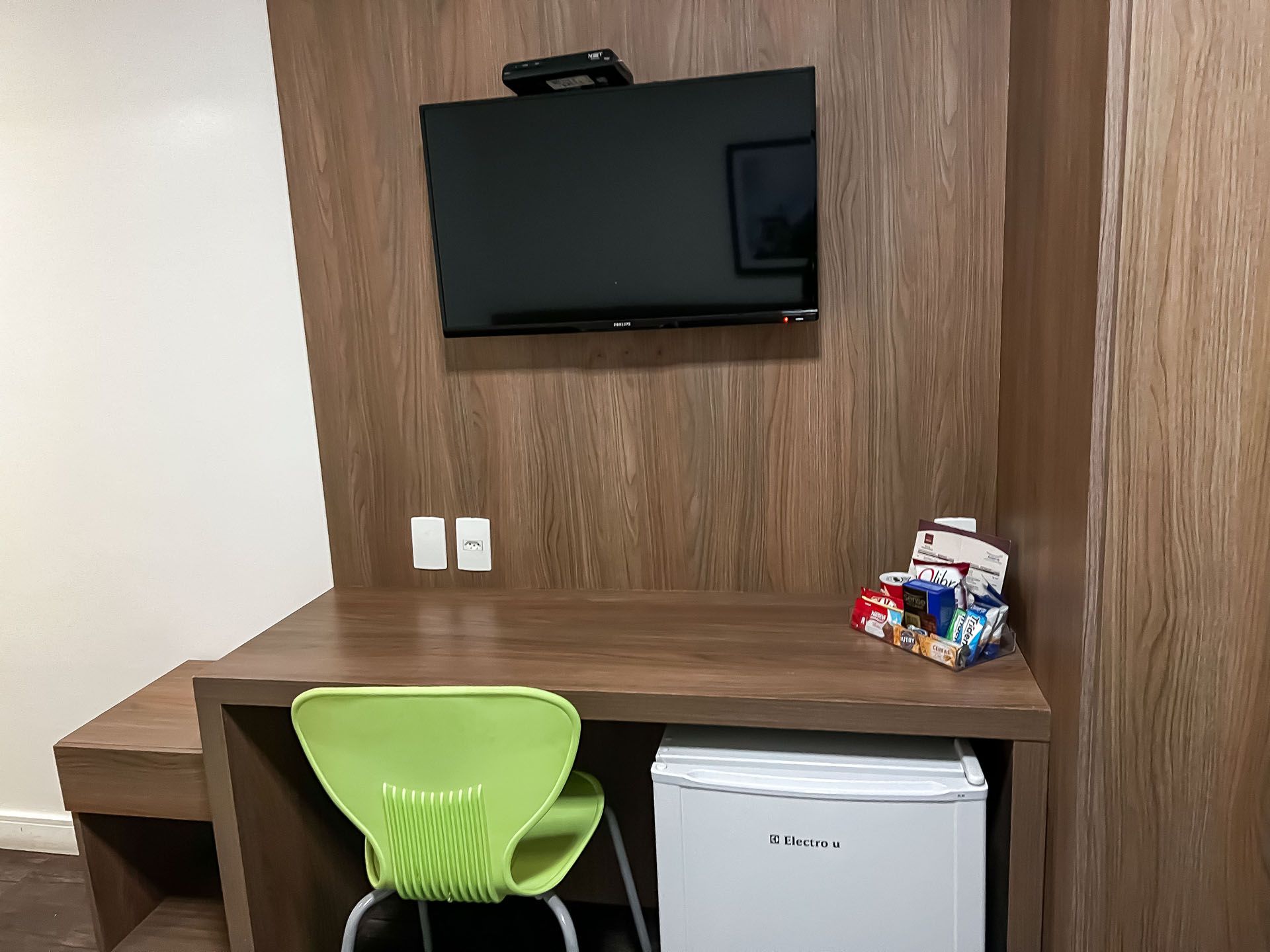 Um pequeno quarto de hotel com TV, escrivaninha, frigobar e uma cadeira verde.