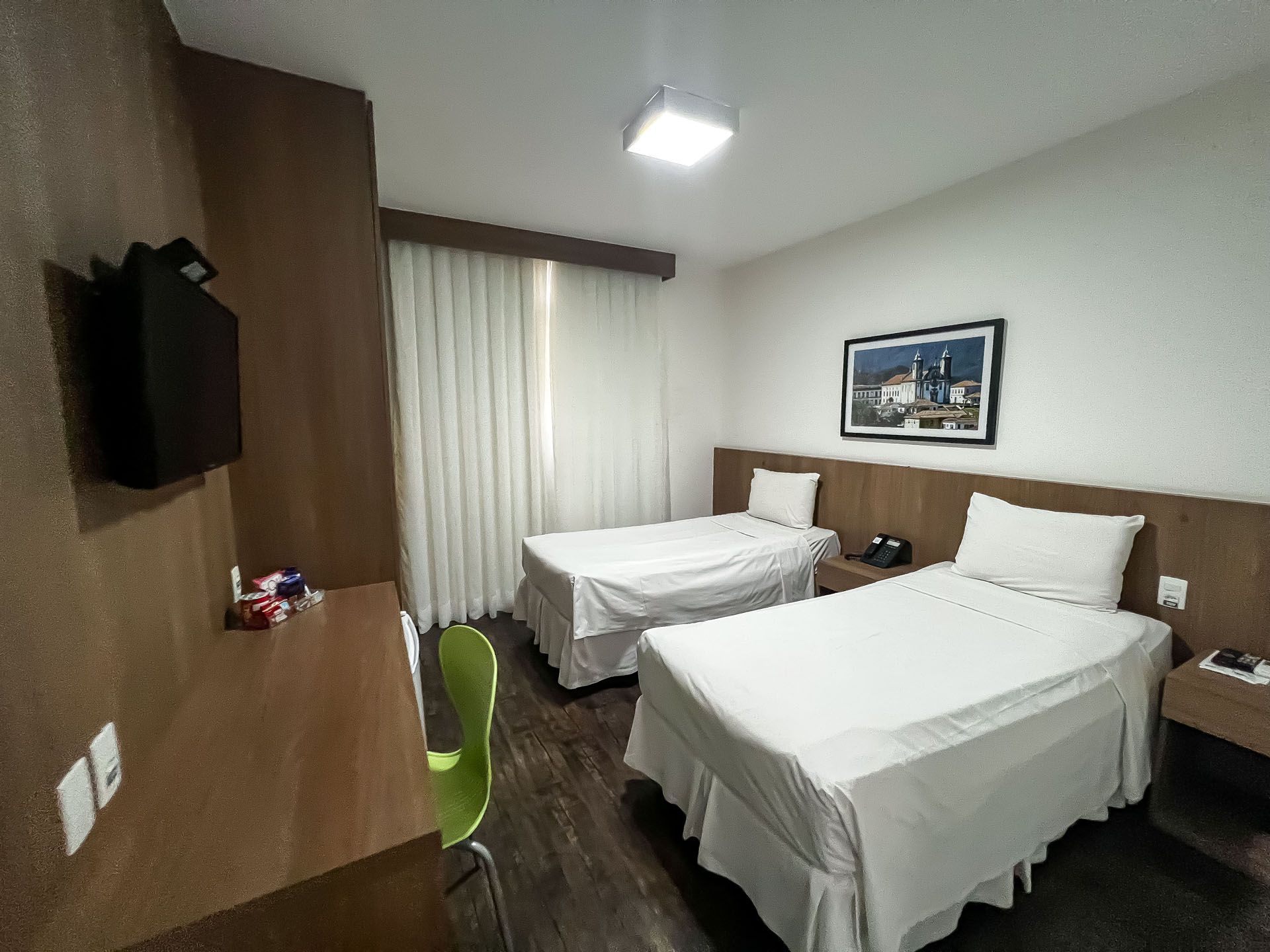 Um quarto de hotel com duas camas de solteiro, uma TV e uma escrivaninha. Roupa de cama branca, piso de madeira e paleta de cores neutras.