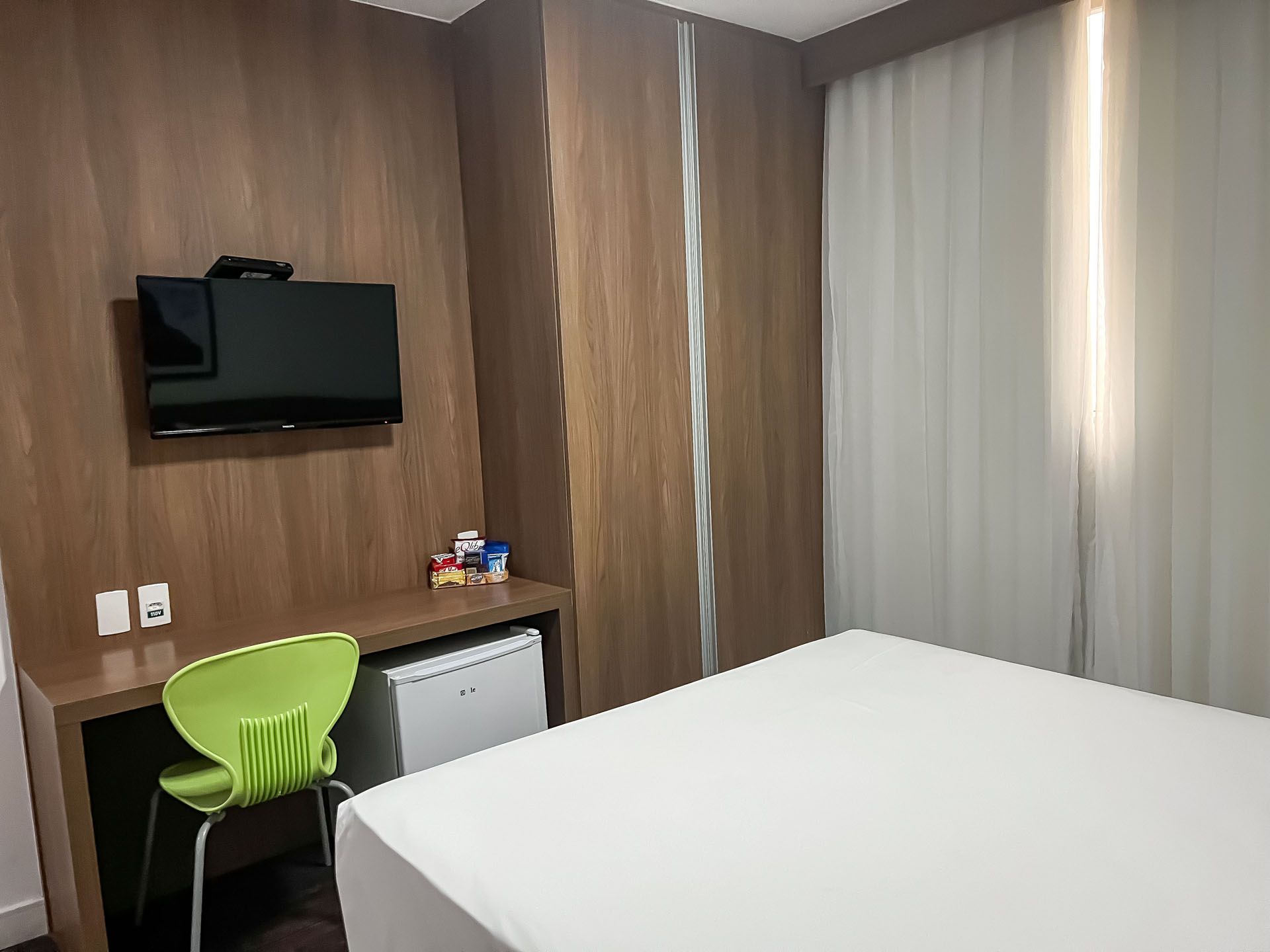 Interior de quarto de hotel com cama, TV na parede, escrivaninha, cadeira verde e frigobar.