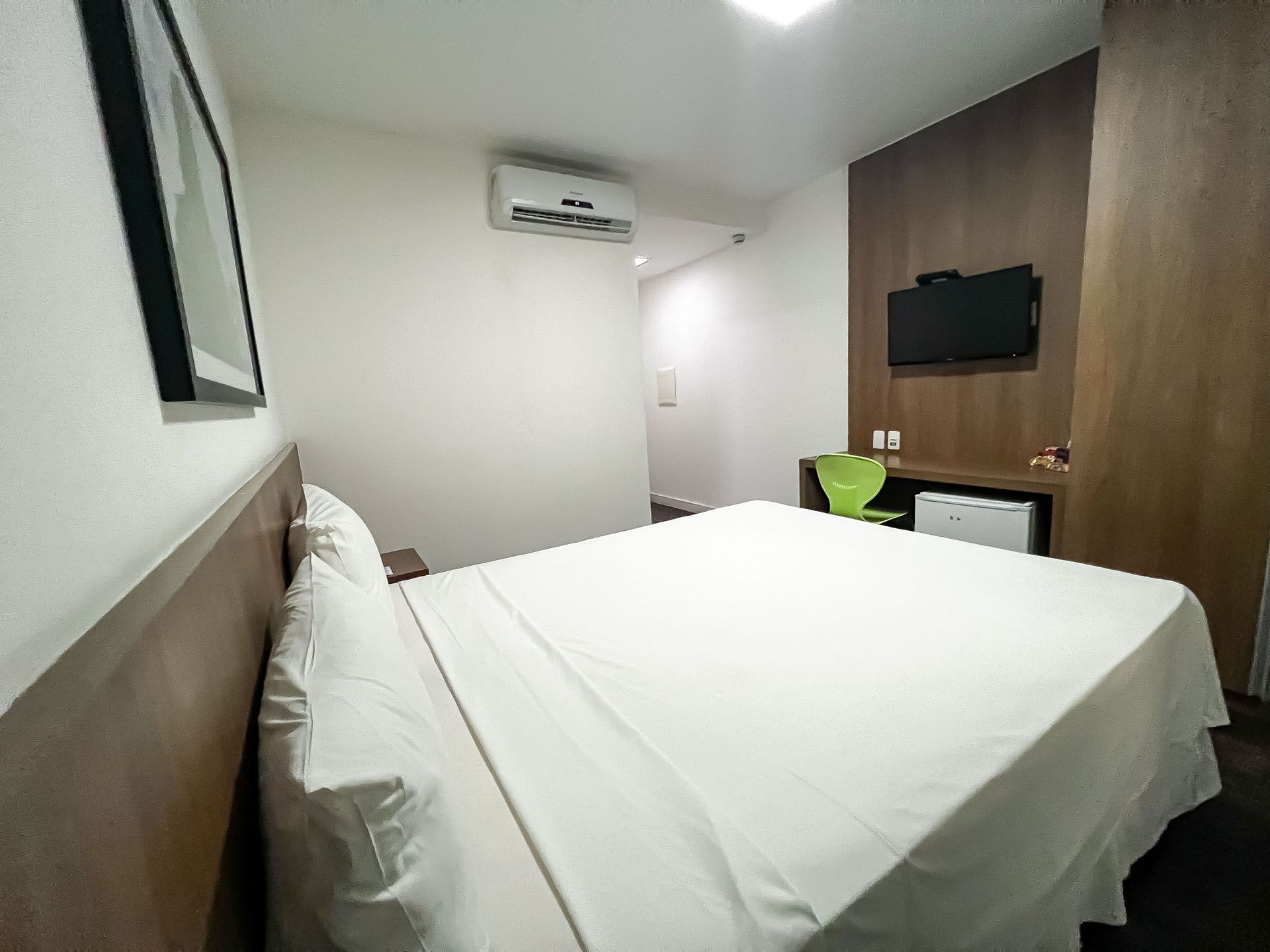 Quarto de hotel com cama, TV, frigobar e uma pequena cadeira verde.
