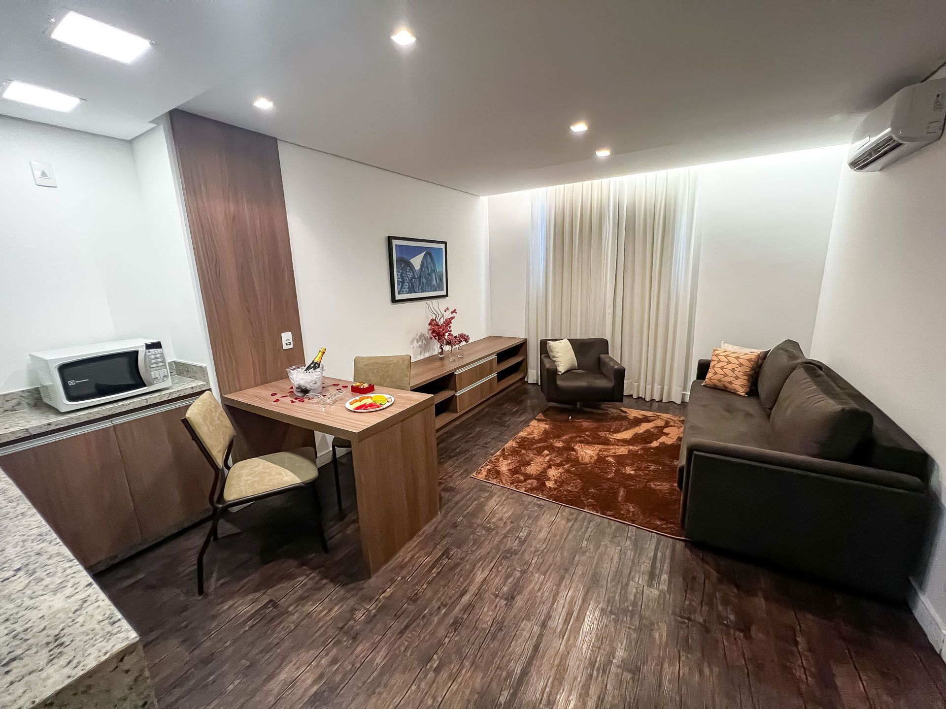 Sala de estar aconchegante com piso de madeira escura, sofá marrom e uma pequena área de cozinha.