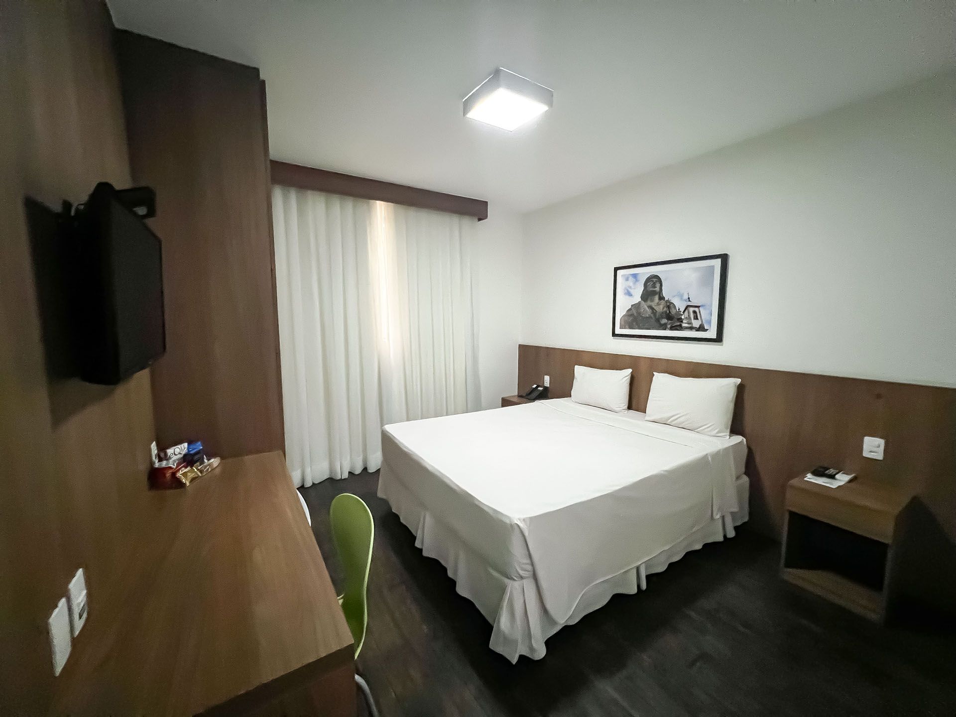 Quarto de hotel com cama, escrivaninha e TV. Paredes e cortinas em tons claros; detalhes em madeira.