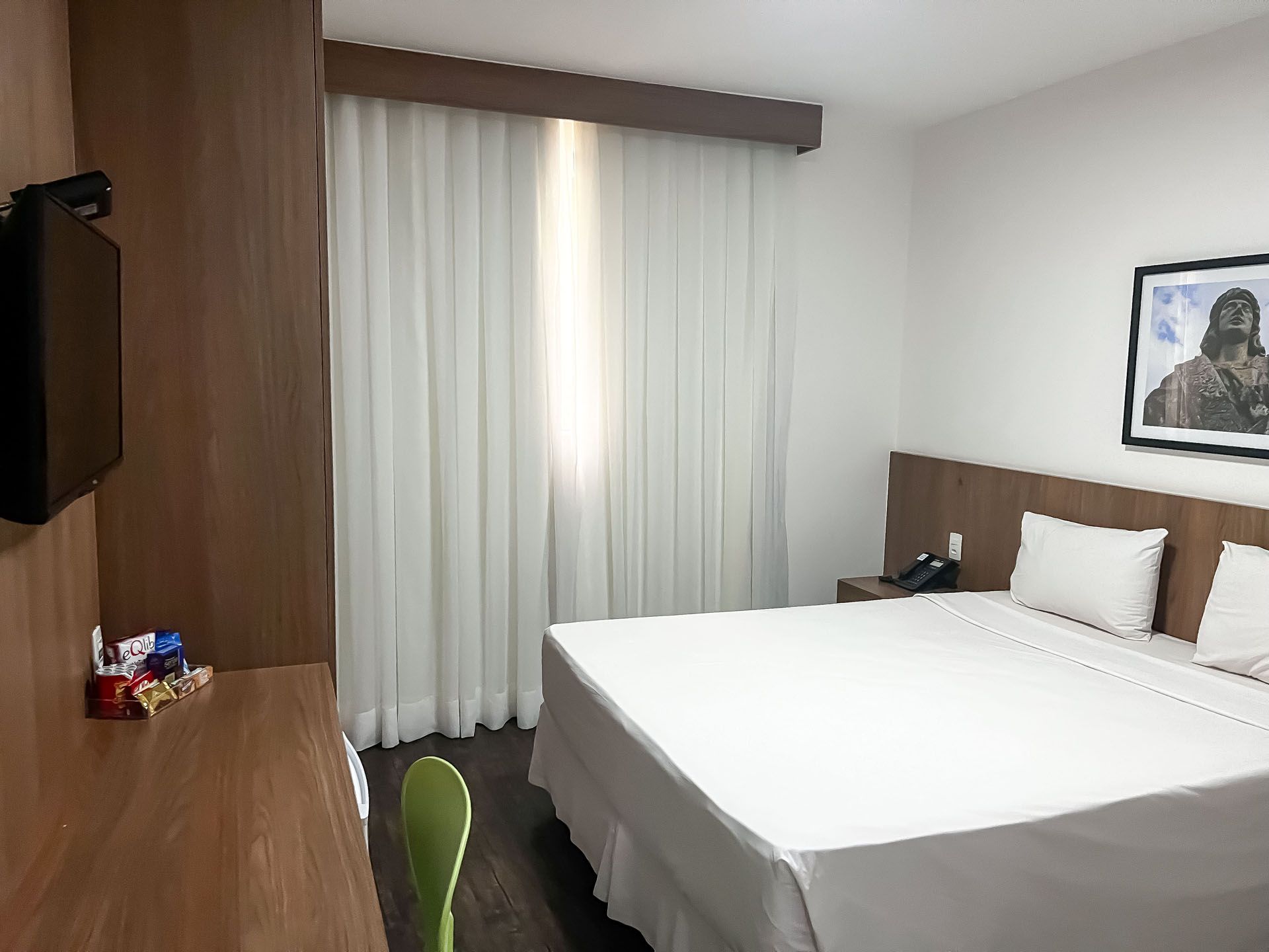 Quarto de hotel com cama, TV, escrivaninha e cortinas.