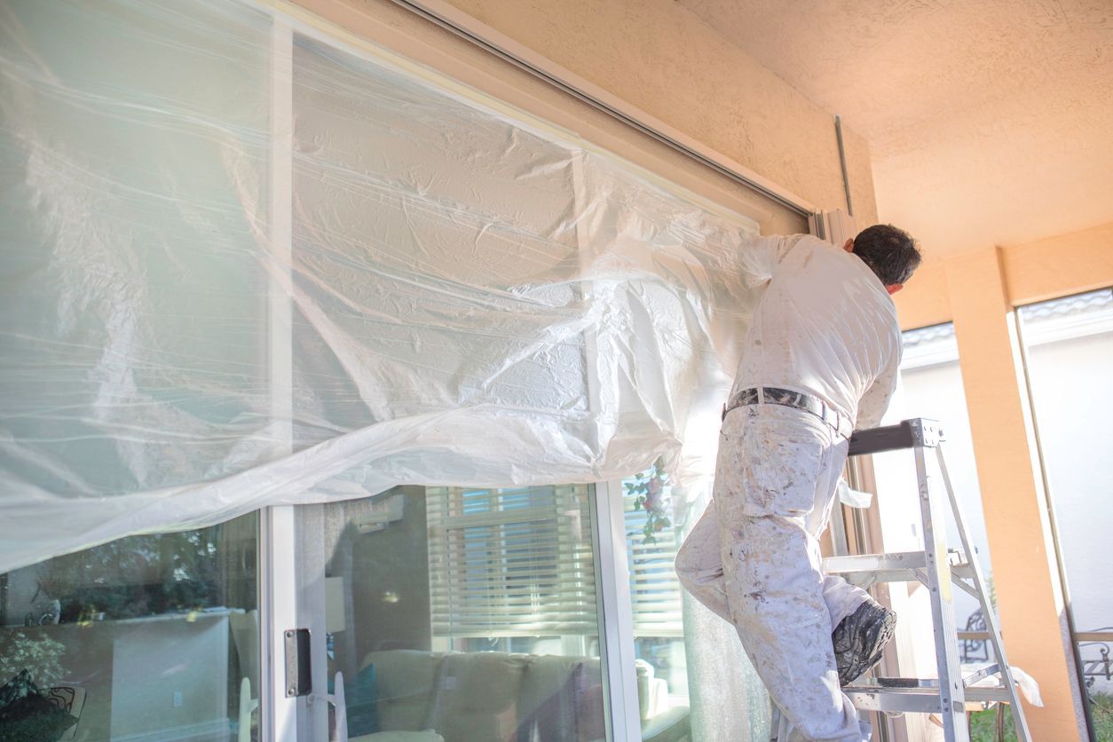 A man is standing on a ladder covering a window with plastic.