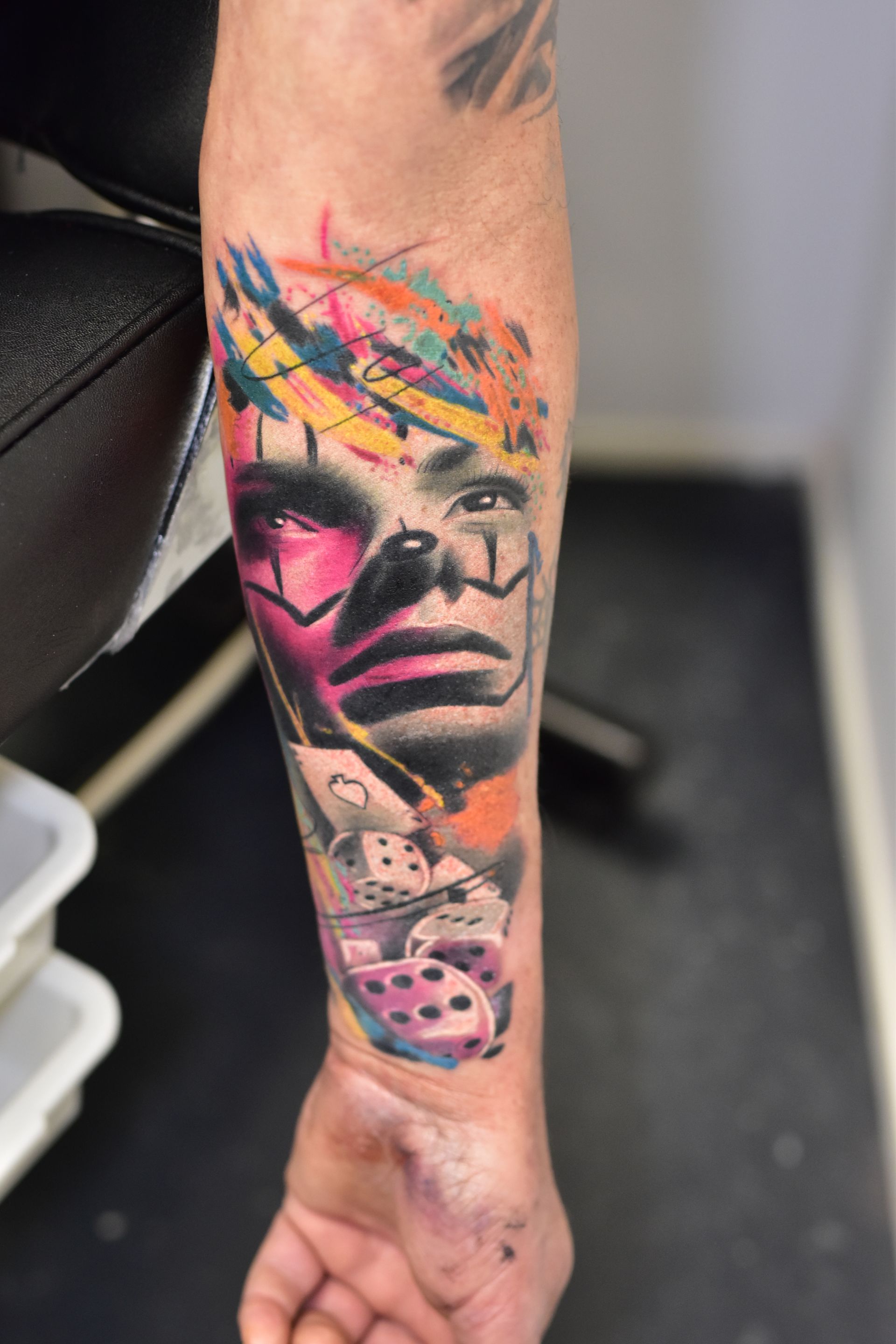 Tatoeage op de arm van een gestileerd gezicht met kleurrijke abstracte elementen en dobbelstenen.